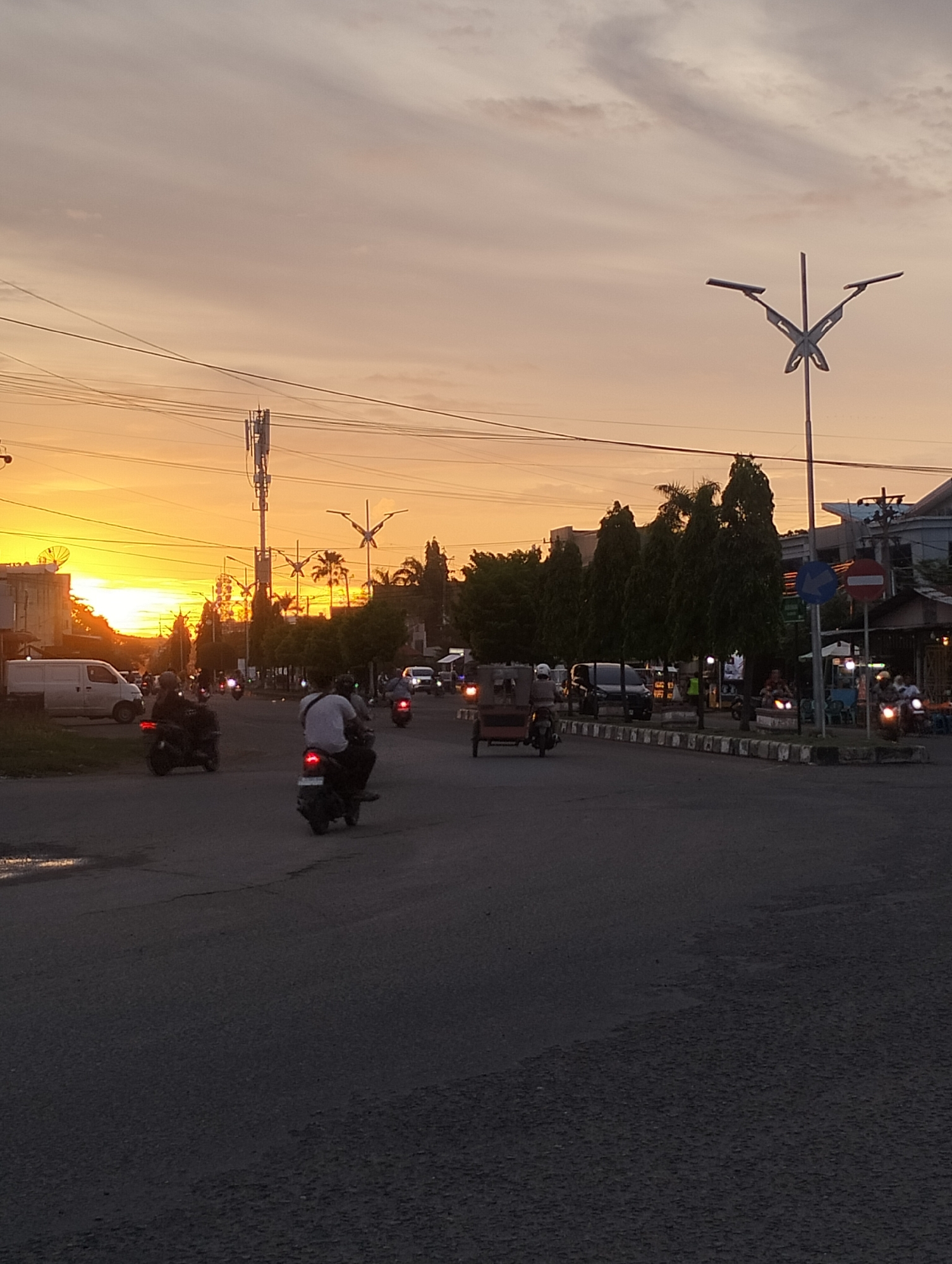 The image depicts a street scene in Lhokseumawe, Indonesia, at sunset. The sky is a gradient of orange and yellow, illuminating the street and surrounding buildings. Several motorcycles are on the road. One motorcycle, driven by a person wearing a white shirt, is in the foreground. A white van is visible on the left side of the road. The street is paved and has a wet patch. The scene includes streetlights with a butterfly design, a cell tower, trees, and a line of buildings in the background.