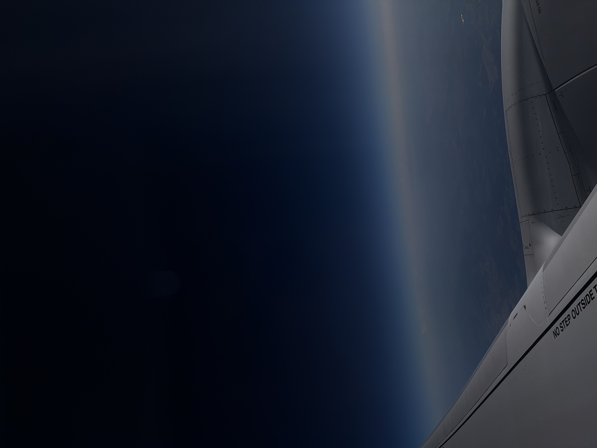 The image shows an aerial view from an airplane window. The upper portion of the image is dark blue, representing the night sky. A thin band of light separates the sky from the Earth below, which appears as a dark landscape with some faint lights. The foreground features a portion of an airplane wing, which is light gray and includes some black lettering that reads "NO STEP OUTSIDE". A section of an airplane engine is also visible on the right side of the image. There are no people or interactions depicted. The location in Wolferborn, Germany is not discernible from the visual information provided in the image.