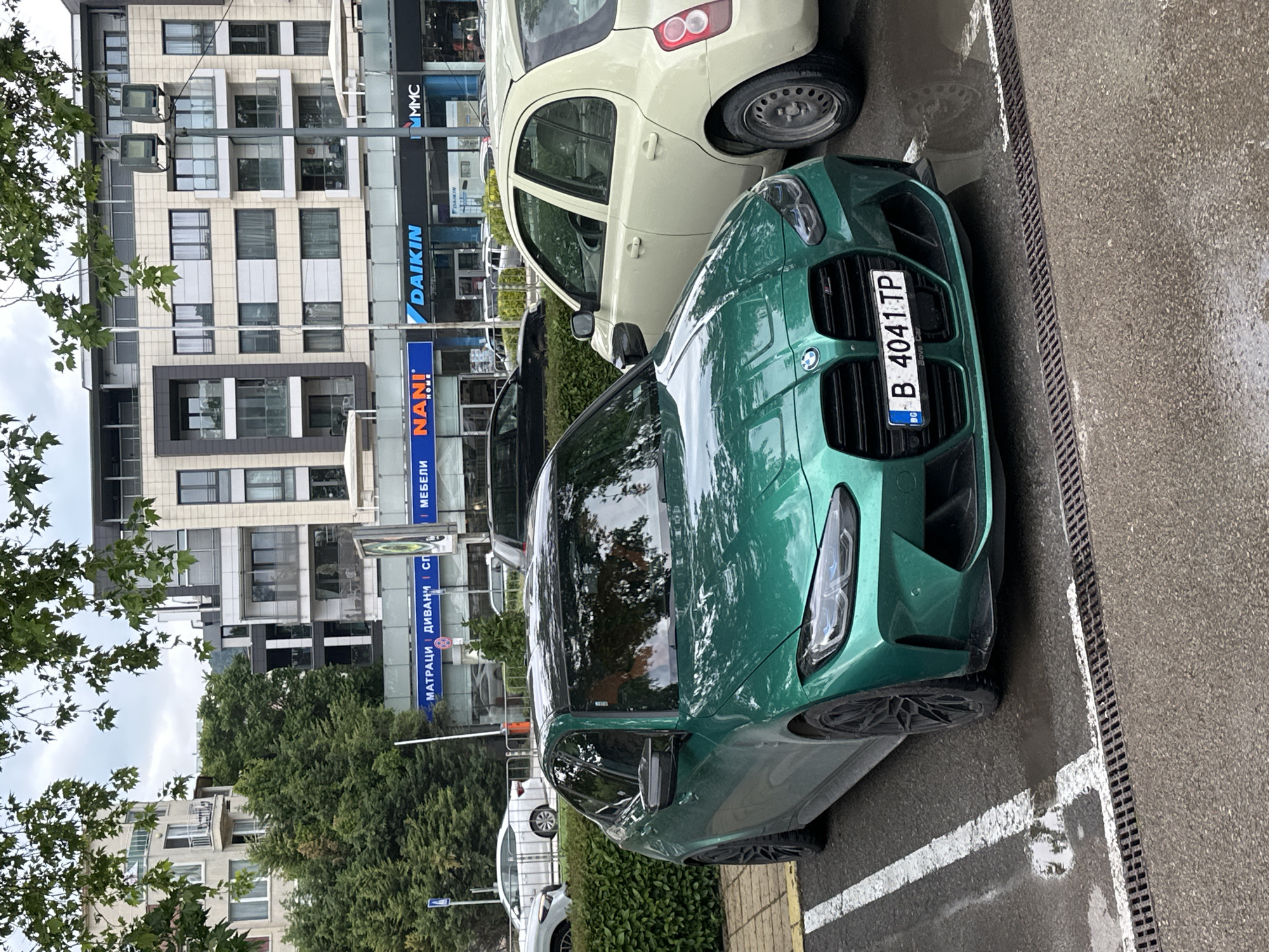 The image shows a green BMW parked on a street in Varna, Bulgaria. The car has a Bulgarian license plate "B 4041 TP". Behind the car, there are other parked vehicles and a building with several signs, including "NANI HOME", "DAIKIN", and signs in Cyrillic script for furniture, mattresses, and sofas. The street appears wet, and there is a tree on the left side of the image. The scene is captured from a slightly elevated perspective.