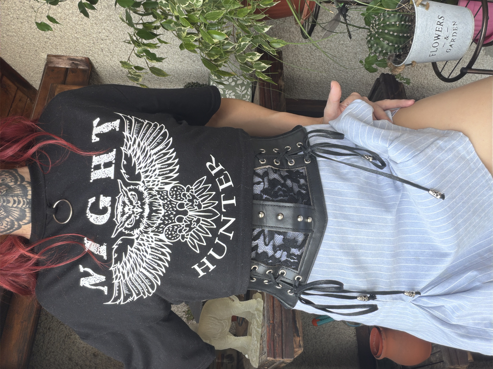 The image shows a person with red hair and tattoos on their neck, wearing a black crop top with an owl graphic and the words "NIGHT HUNTER". They are also wearing a black corset with lace detailing over a light blue pinstripe skirt. The person is standing outdoors in front of a textured wall, with wooden shelves holding a light-colored statue and plants. A cactus in a pot sits on a metal stand to the right. The location is Svilengrad, Bulgaria.