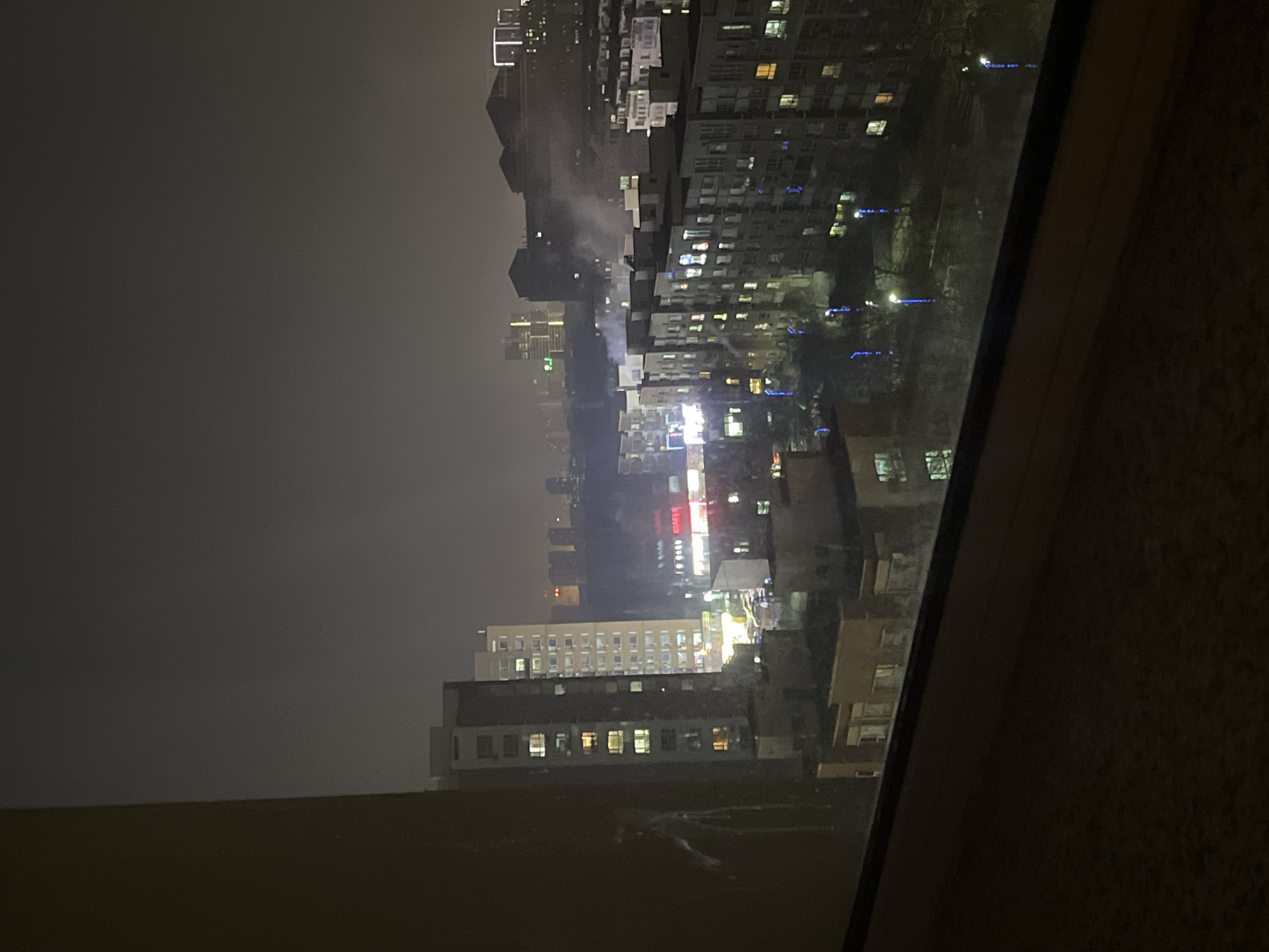 View of Changchun City, China at night from an elevated position, likely through a window. The city is visible with multiple high-rise buildings, illuminated by streetlights and lights from the buildings. There is low visibility due to fog or smog. Some buildings have distinctive lighting features, like the use of blue light. The foreground shows the edge of the window frame.