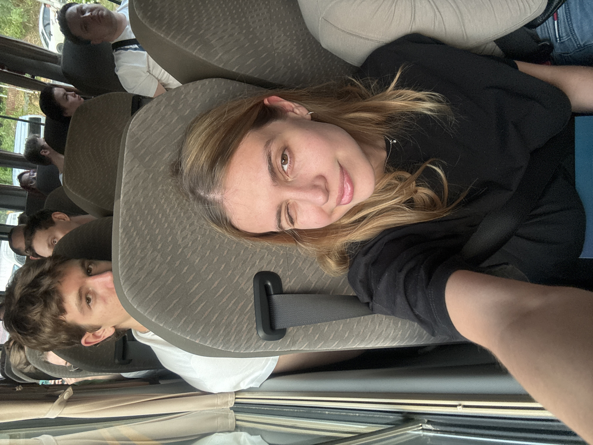 The image shows the interior of a bus filled with passengers. A fair-skinned young woman with blonde hair and a black shirt is in focus, winking at the camera. A fair-skinned young man in a white shirt is partially visible behind her seat. Other passengers are visible in the background, some appearing to be sleeping or resting. The seats are upholstered in a neutral gray fabric. A seatbelt is visible across the woman's seat. The environment suggests a vehicle interior, likely a tour bus, filled with passengers. The city of Patong, Thailand cannot be confirmed.