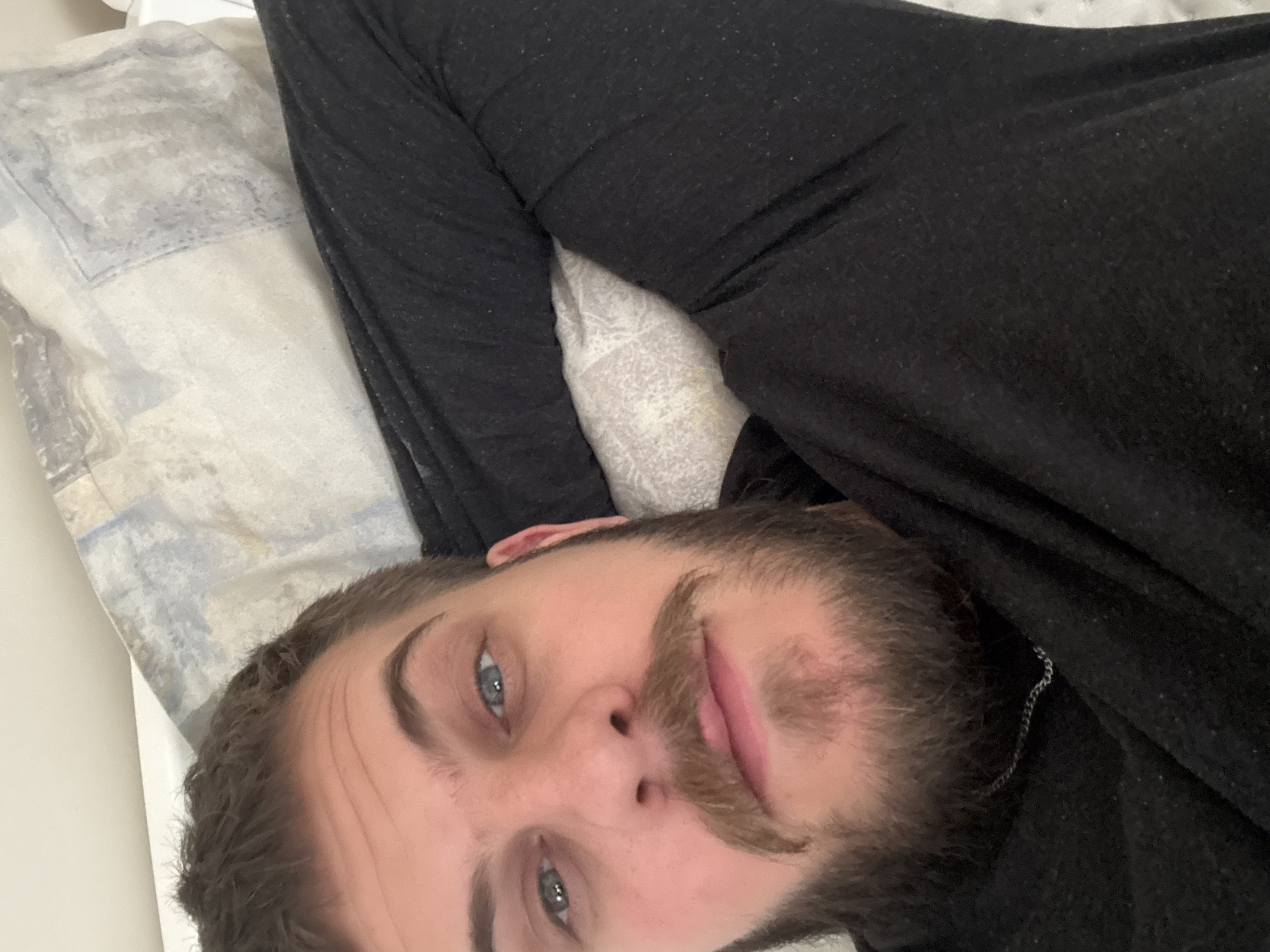 A man with light blue eyes, brown hair, and a beard is lying on his side on a bed. He is wearing a black long-sleeved shirt. The pillow has a light blue and white pattern. A silver chain is visible near his neck. The image's location cannot be determined from the information provided.