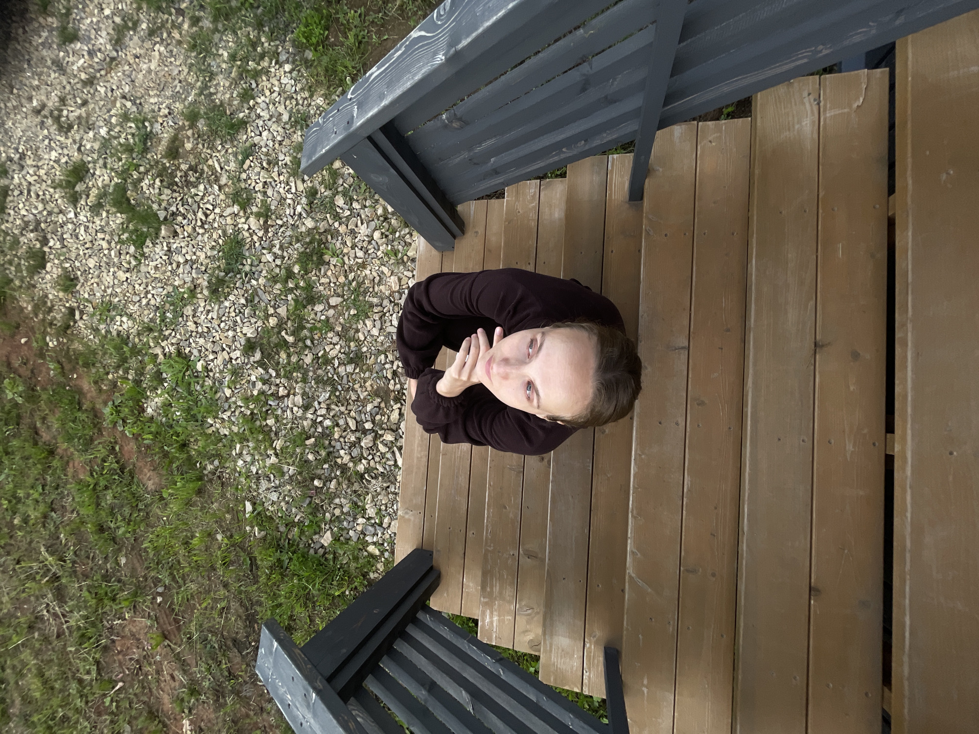 A high-angle shot captures a woman seated on a wooden staircase, looking upwards. She has fair skin, light brown hair pulled back, and is wearing a dark brown long-sleeved shirt. Her arms are crossed in front of her chest, and her hands are resting near her chin. The woman is positioned on the middle steps of the staircase, which is flanked by dark gray railings. The staircase leads down to an area with brown gravel and patches of green grass. The background shows more grass and a dirt path. The location is identified as Podshivalovo, Russia.