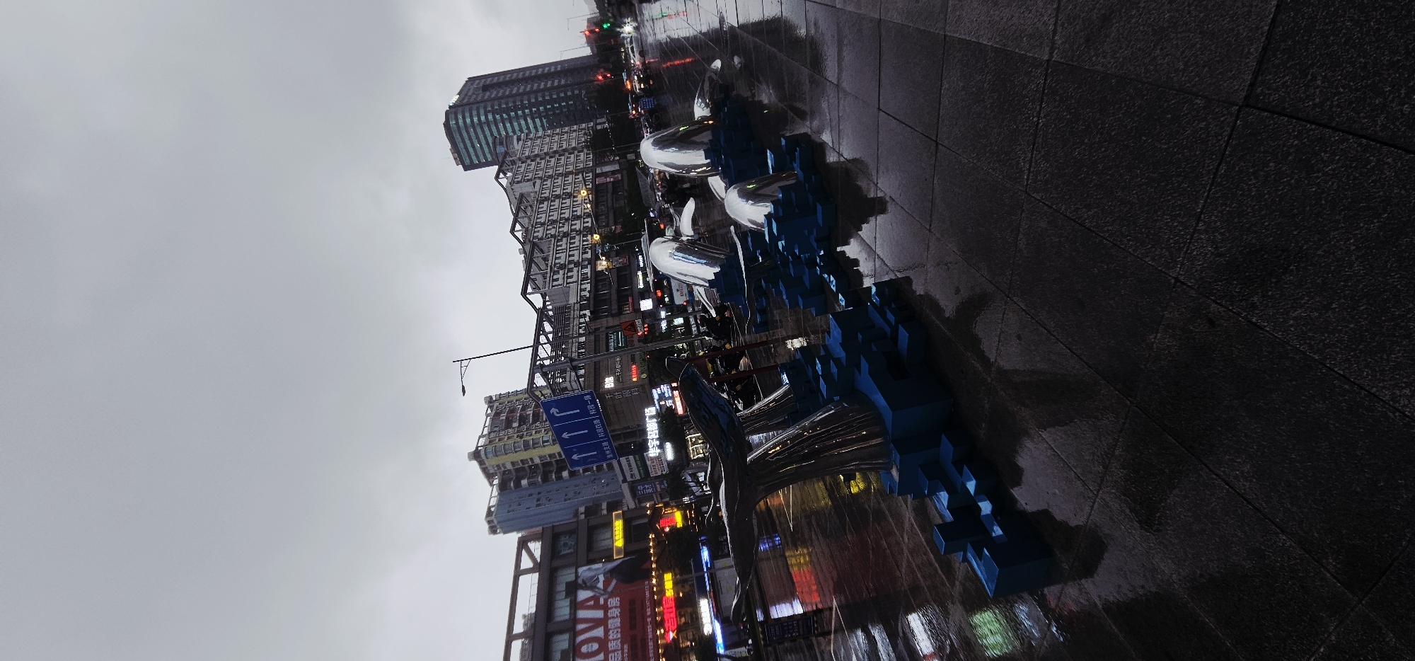 This image depicts an urban street scene in Jiulongpo District, Chongqing, China, during twilight or early evening under overcast conditions. The ground, paved with dark square tiles, is visibly wet and highly reflective, suggesting recent rain.
In the foreground and mid-ground, a public art installation is prominent. It consists of multiple metallic, highly reflective sculptures resembling whale tails and bodies emerging from a base of interconnected blue, pixelated rectangular prisms. At least three distinct chrome-like whale tail sections are visible, reflecting distorted ambient light and colors.
The background features several multi-story buildings lining the street. On the left, a commercial building displays a large illuminated sign with "NOVA" and Chinese characters that include "健身房" (gym). In the center, a blue street sign with white arrows (indicating straight, left, and right turns) prominently displays Chinese characters, including "渝州路" (Yuzhou Road) and "科技园一路" (Science Park Road 1), confirming the location within Jiulongpo District. A black street lamp post stands near this sign. Further to the right, a tall, dark skyscraper with green-tinted windows is visible, alongside other buildings with various illuminated signs in Chinese.
Distant street traffic is indicated by blurred vehicle shapes and illuminated headlights/taillights, along with visible traffic lights showing red and green. A few indistinct human figures are discernible in the mid-ground, likely pedestrians. The sky is a uniform, light grey, indicating heavy cloud cover. The entire scene is captured from a slightly low and tilted perspective.