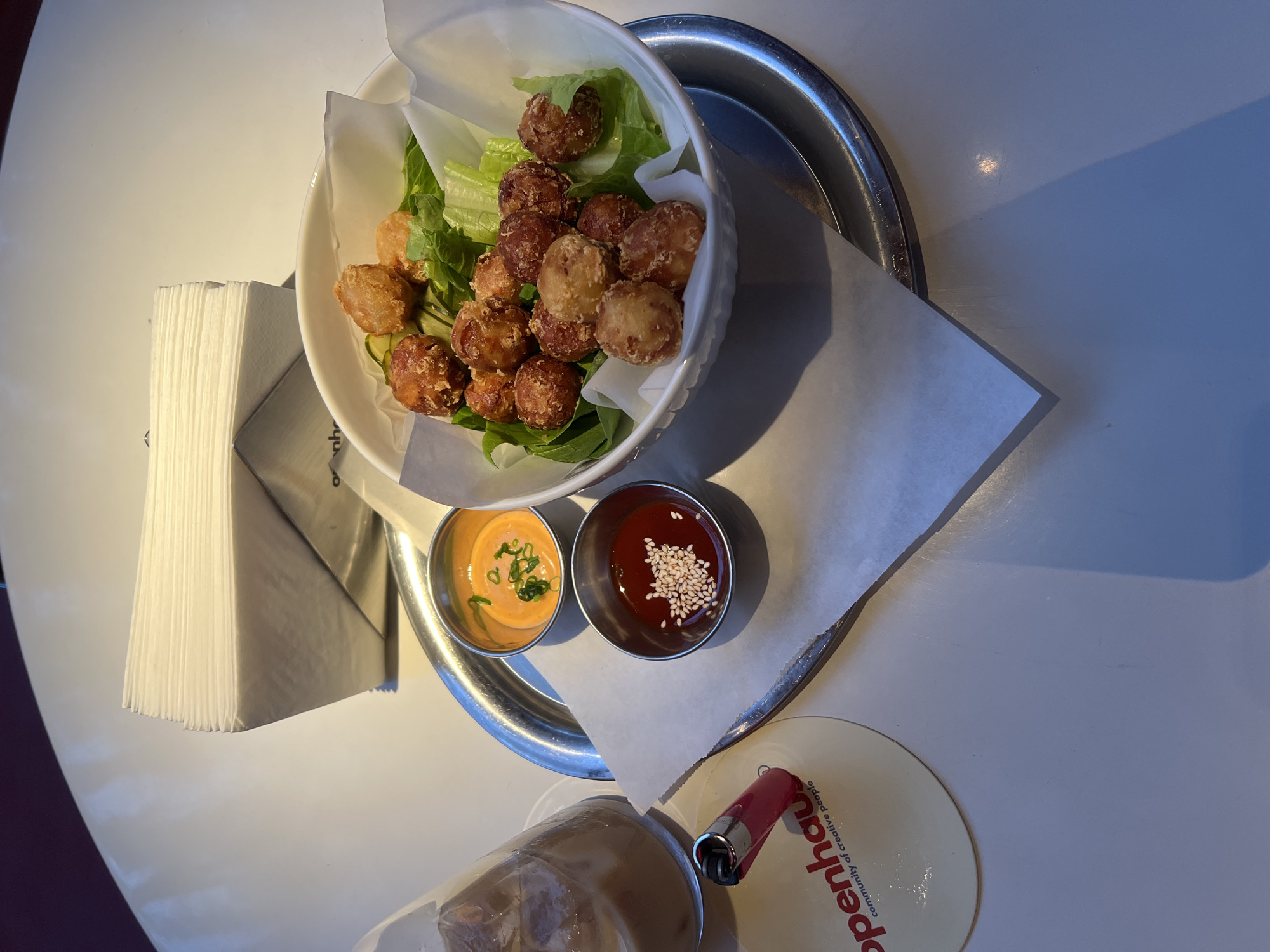 The image depicts a meal served on a silver tray. The main dish consists of fried balls served on a bed of lettuce in a white bowl lined with parchment paper. Two small metal bowls contain sauces, one with a creamy orange sauce garnished with green onions and the other a red sauce topped with sesame seeds. A stack of napkins is placed next to the tray. In the lower portion of the image is a glass with a drink and a red lighter next to a coaster with the word "openhau." The coaster suggests the image was taken at Openhau, a community of creative people, which has a location in Yerevan, Armenia. The table is white, and the lighting suggests an indoor setting.