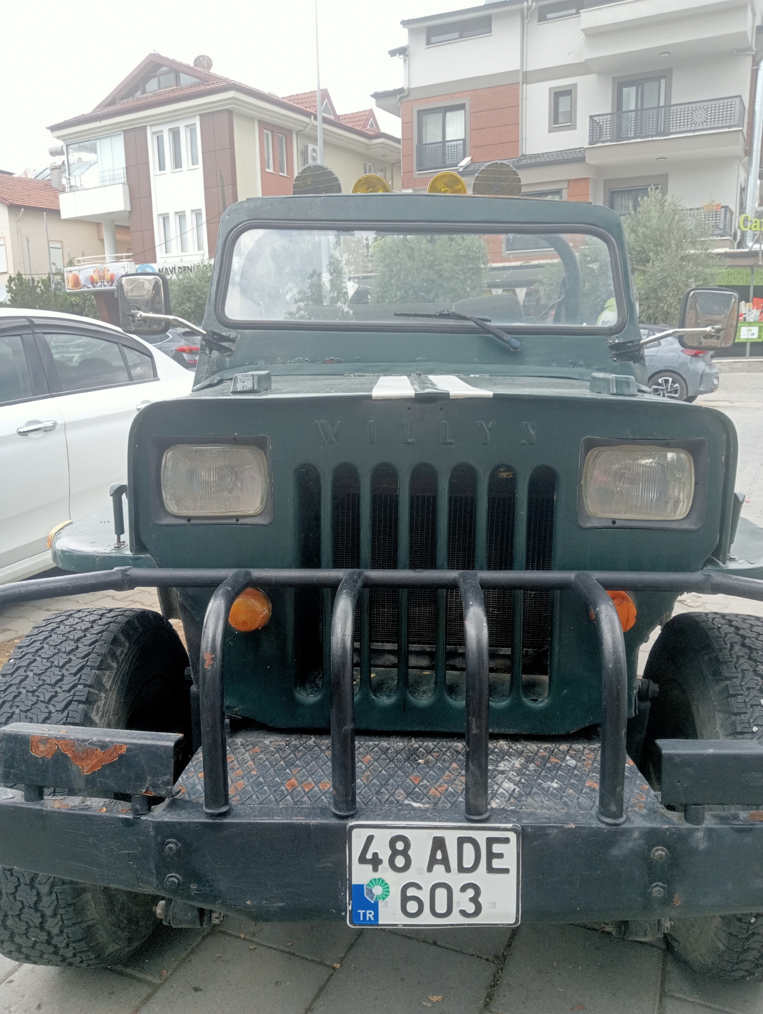 An olive green Willys jeep is parked on a paved street in Fethiye, Turkey. The jeep has a black bumper, prominent headlights, and a grille with vertical slats. A license plate reading "48 ADE 603 TR" is affixed to the front bumper. Behind the jeep, a white car is visible, as well as a multi-story building with a reddish-brown and white facade. The sky is overcast.