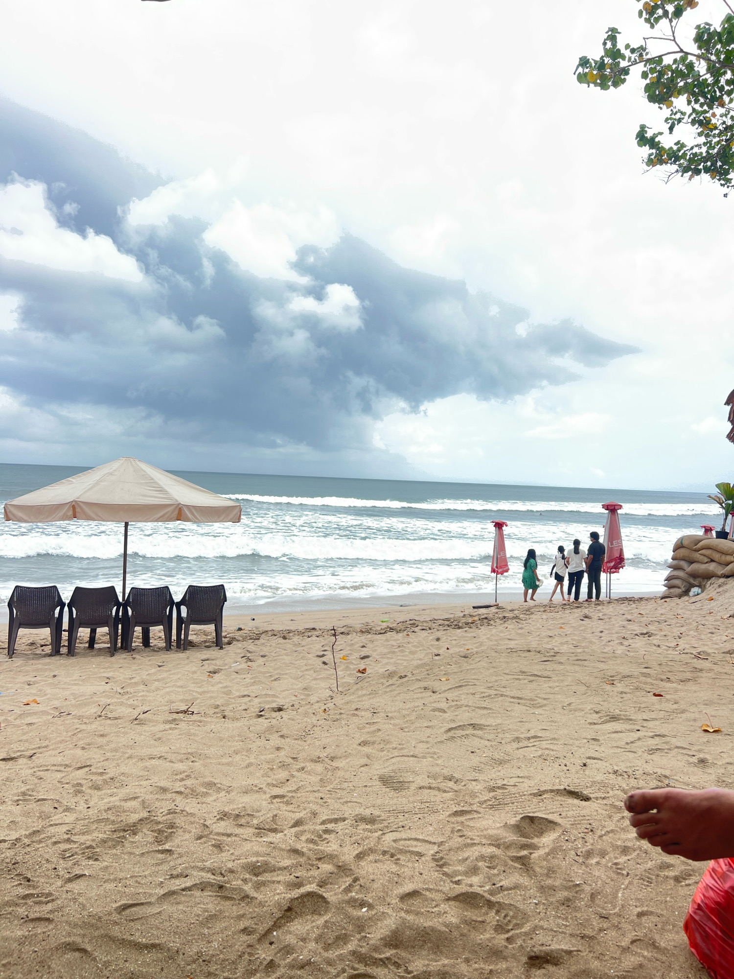 The image depicts a beach scene in Kuta, Indonesia, under a cloudy sky. In the foreground, the sandy beach is visible with footprints. To the left, four dark plastic chairs are arranged under a beige beach umbrella. Further back, on the sand, a group of four people are standing, facing the ocean. There are also two red umbrellas with white lettering visible behind them. The ocean, with moderate waves, stretches to the horizon. In the far right of the image, sandbags are stacked, and a person's foot is visible in the lower right corner. The environment appears to be a coastal area with vegetation visible in the upper right portion of the frame.