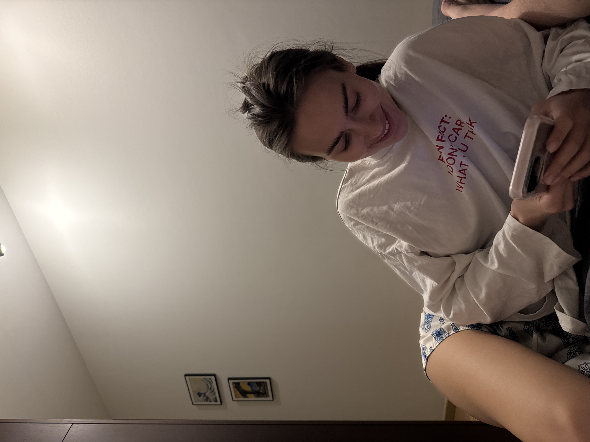 A young woman is holding a cell phone, wearing patterned shorts and a white long-sleeved shirt that reads "I don't care what u think". Two framed pictures are hanging on a wall in the background. The city and location are unknown.