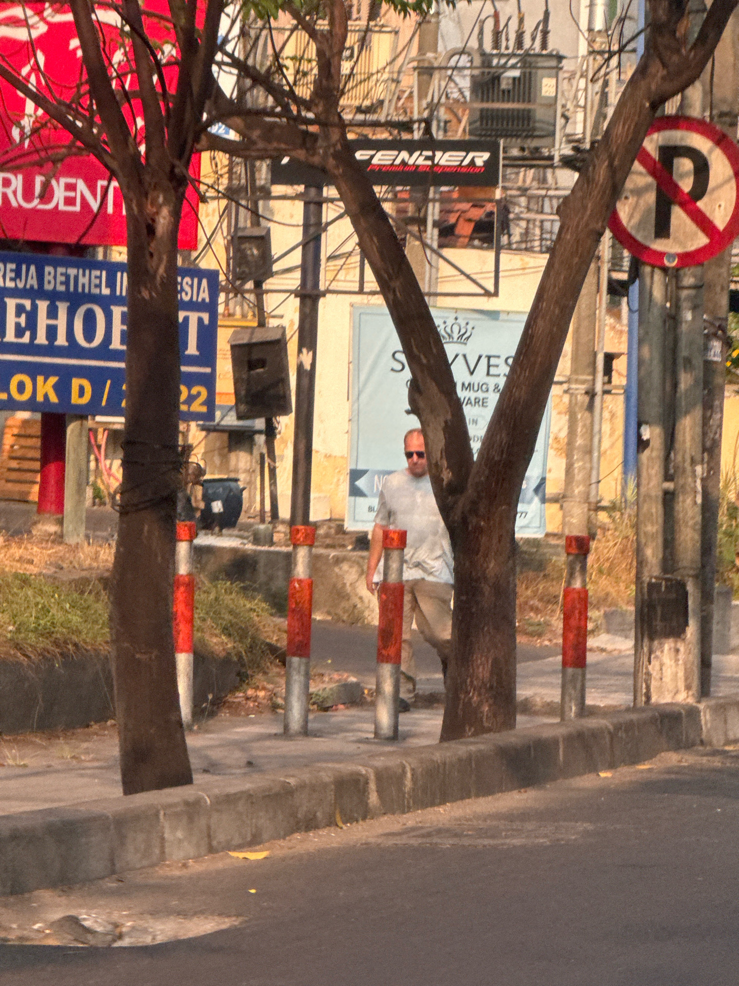 A man wearing a light-colored short-sleeved shirt and light-colored pants walks on a sidewalk. He is partially obscured by tree trunks. The sidewalk is lined with concrete bollards painted with red and white stripes. Behind the bollards and sidewalk is a patch of dry grass and dirt. In the background, there are several trees with thin trunks and branches, some bearing leaves. There are also street signs, electrical wires, and poles. One sign is circular with a red border and a red diagonal line over a black letter 'P', indicating no parking. Another sign is rectangular with red text on a black background that appears to read "FENDER". There are also other signs with blue and white text. The ground is paved.