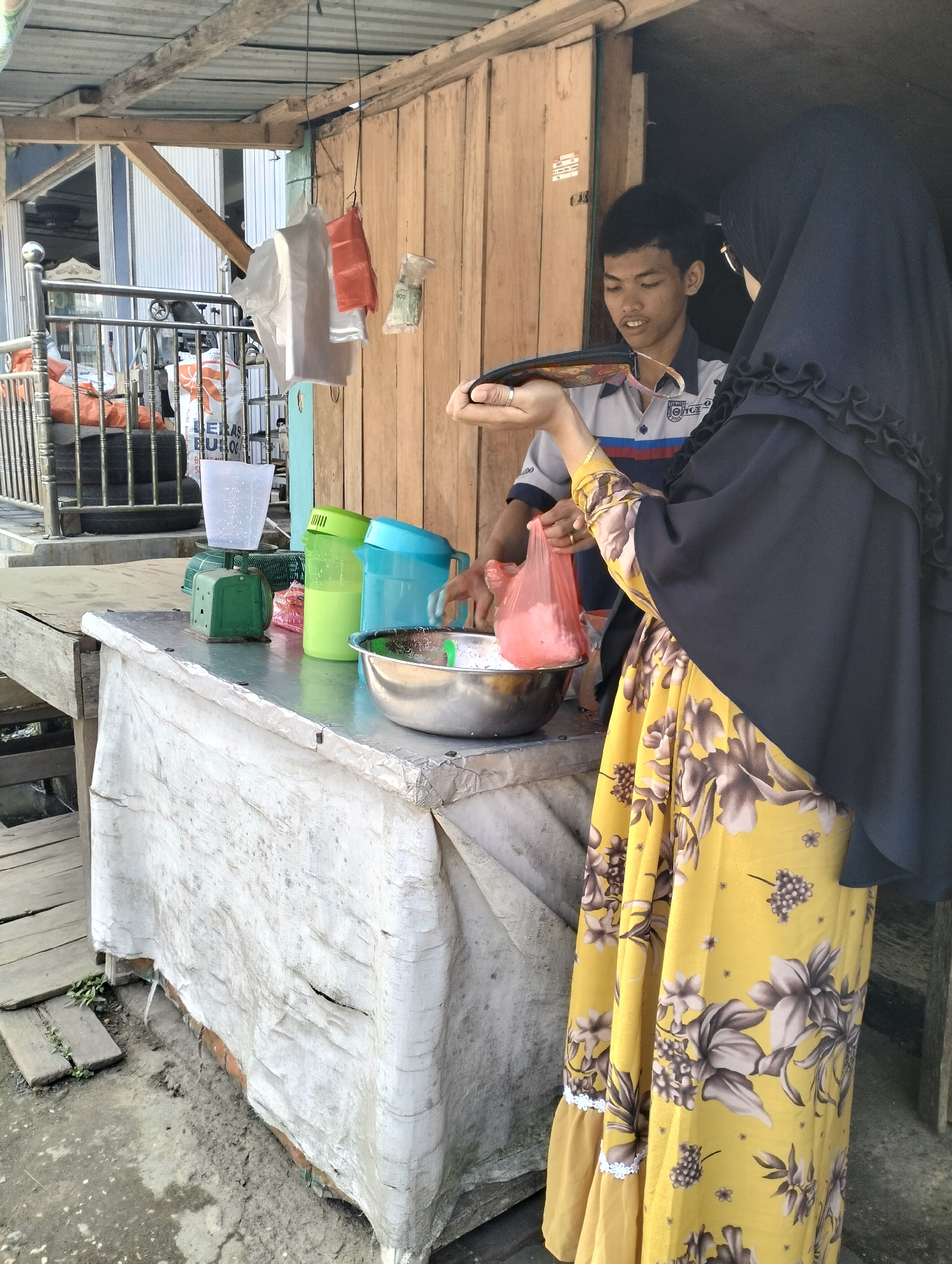 In Pematang Lumut, Indonesia, this scene depicts a market stall. A young man, wearing a striped shirt, stands behind a counter. A woman, draped in a black hijab and a vibrant yellow floral dress, is interacting with him. She holds a pink plastic bag containing a light-colored substance, and he appears to be reaching for it or observing her transaction. On the counter, there are various items: a green weighing scale with a clear measuring cup, two colorful jugs (one lime green, one blue), and a silver bowl. A white tarp covers the front of the stall. In the background, there's a wooden structure and a metal railing with bags stacked behind it. The ground is dirt and concrete.