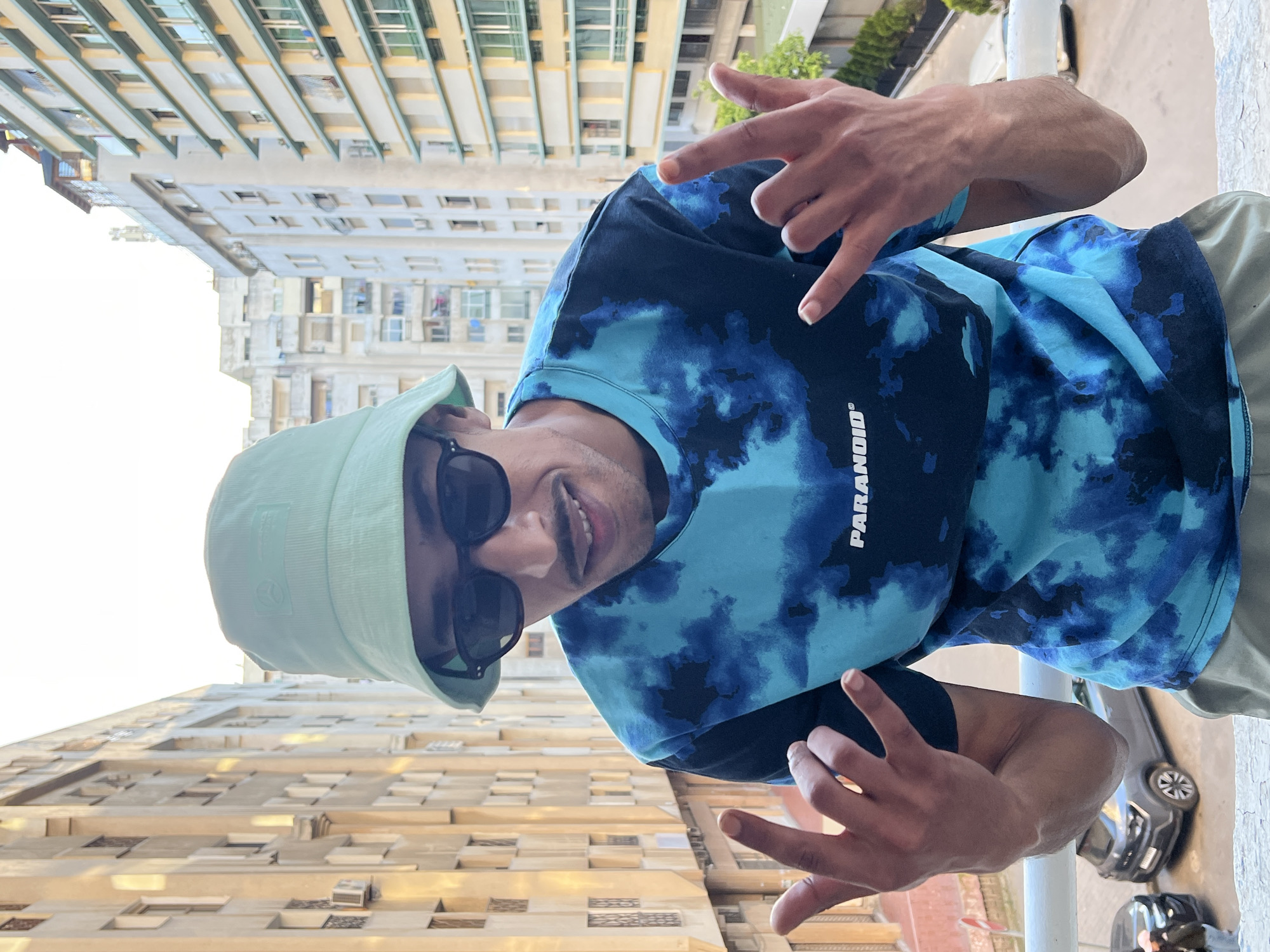 A man stands on a balcony, wearing a light green bucket hat and sunglasses. He is also wearing a blue tie-dye t-shirt with the word "PARANOID" printed on it. He is holding up both hands, making hand gestures with his fingers. Behind him are tall apartment buildings, suggesting an urban setting. A car is visible on the street below. The location appears to be the city of Tanger طنجة, Morocco.