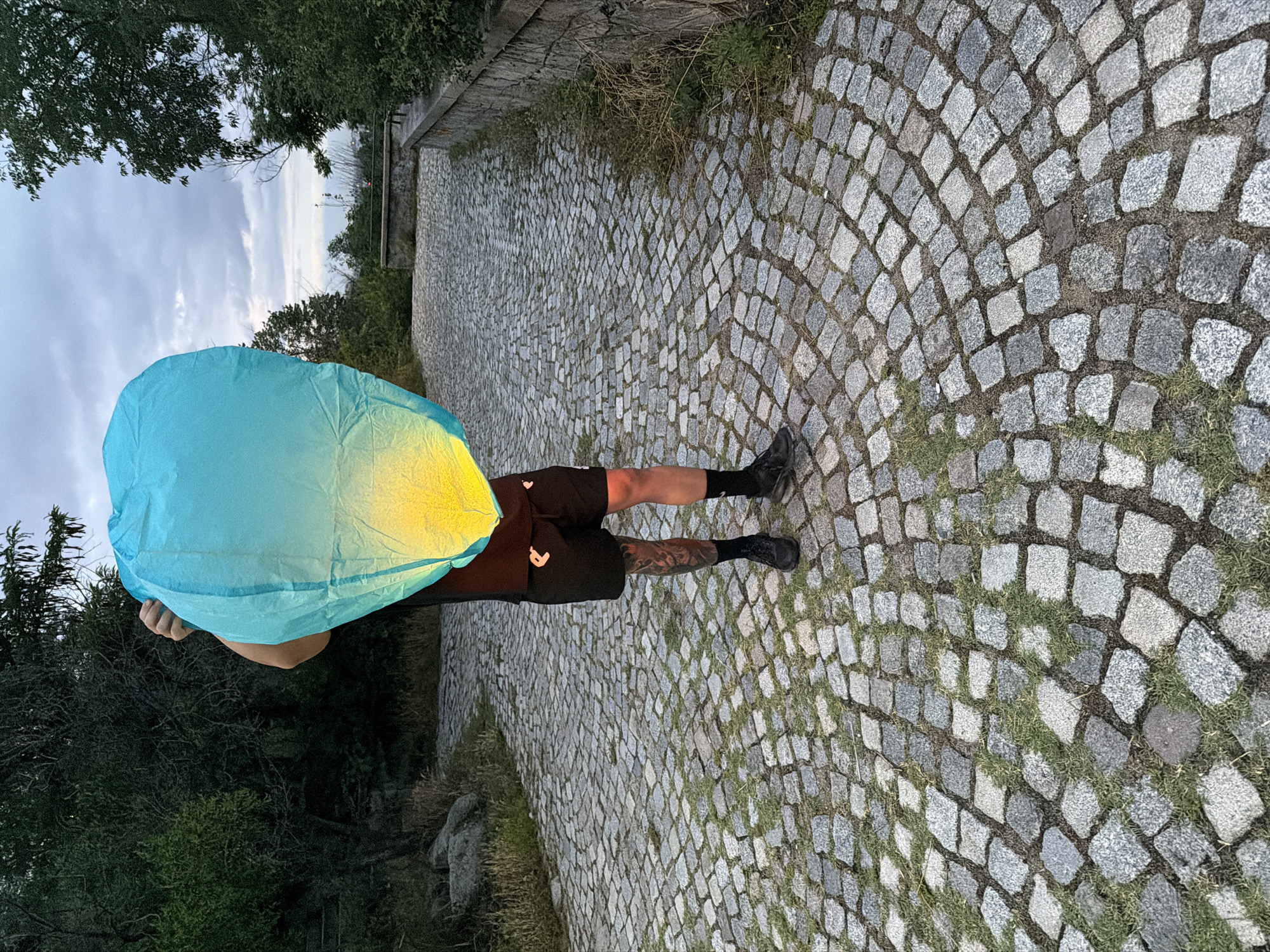 A person stands on a cobblestone pathway, holding a large, unlit sky lantern which is predominantly blue with a yellow gradient towards the bottom. The person is wearing black shorts and a black t-shirt, with visible tattoos on their left leg. They are obscured by the lantern, with only their legs and lower torso visible. The pathway is bordered by grass and a stone wall on the right. Trees and vegetation are present on both sides of the pathway, and the sky above is overcast. The scene appears to be an outdoor location, possibly a park or a viewpoint. Based on the architecture of the stone wall and the surrounding vegetation, this location strongly resembles the area around the Alyosha Monument (Monument to the Soviet Army) in Plovdiv, Bulgaria, specifically along one of the scenic viewpoints or paths leading to it.
