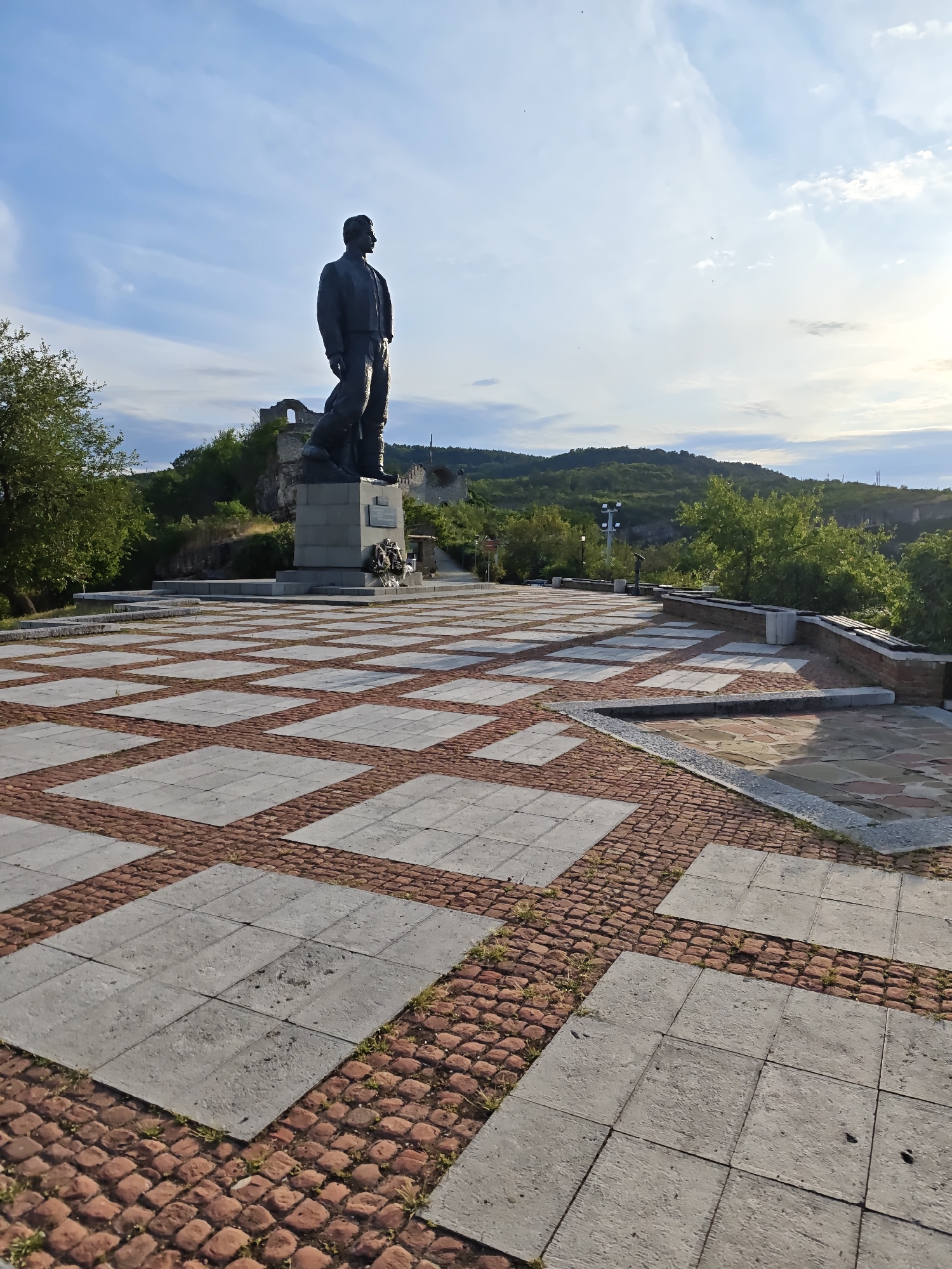 The image depicts a wide-angle view of a paved plaza featuring a large bronze statue of a man standing on a pedestal. The plaza's paving is a patterned design of large, light-colored square tiles interspersed with smaller, reddish-brown brick-like stones arranged in a diagonal grid. In the background, there are remnants of stone structures, likely ruins, situated on a hillside covered with greenery. Further back, rolling hills covered in trees form the horizon under a bright, partly cloudy sky. This scene is located at the Monument of Vasil Levski in Lovech, Bulgaria.