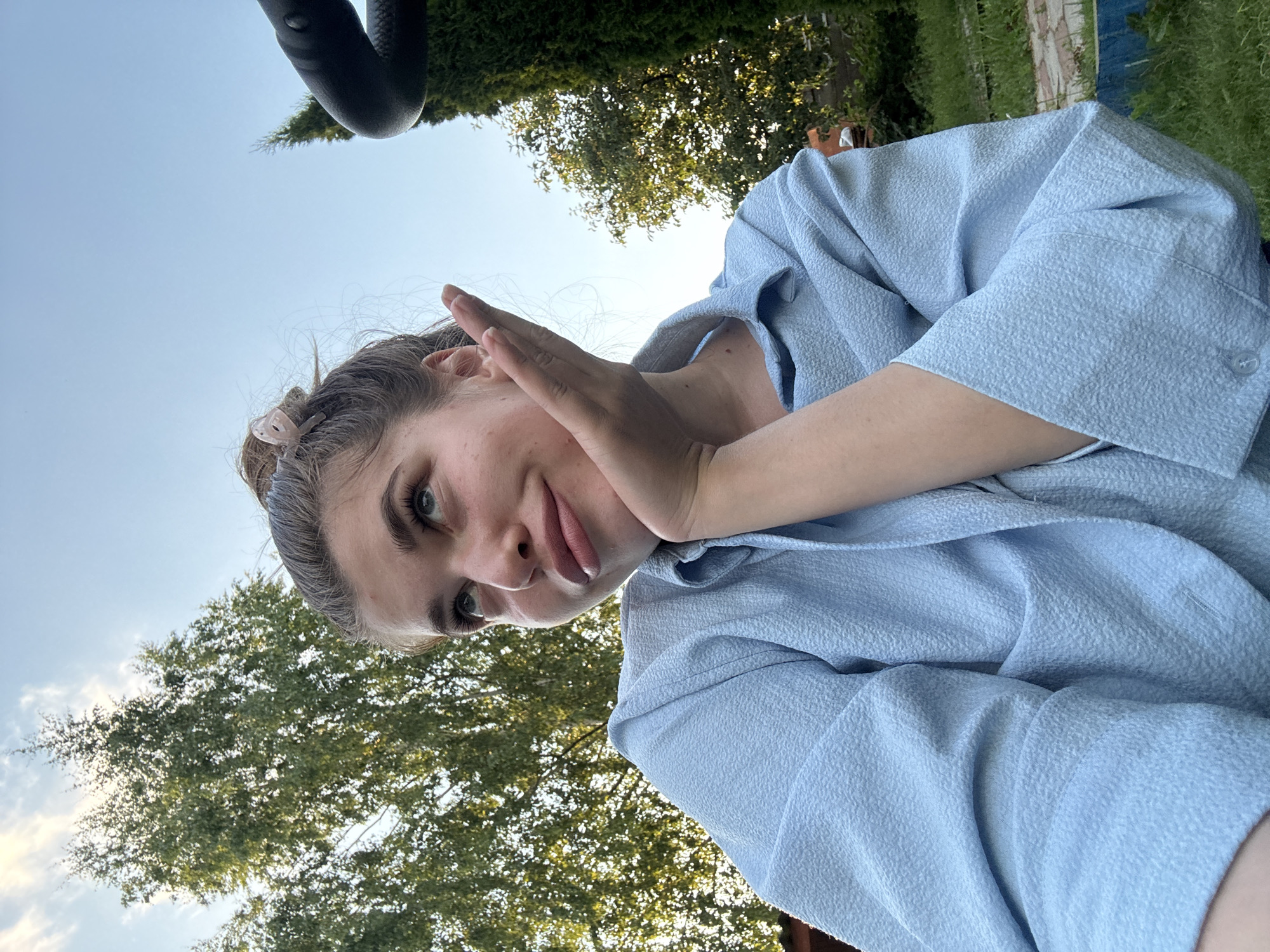 The image shows a young woman with her hair pulled back, wearing a light blue textured shirt. She is holding her hand up to her face, obscuring part of it, and looking towards the camera with a playful expression. The background consists of green trees and a clear sky, suggesting an outdoor setting during the daytime. The image is oriented sideways. There is no visible text. The location context provided is Мещёры, Russia.