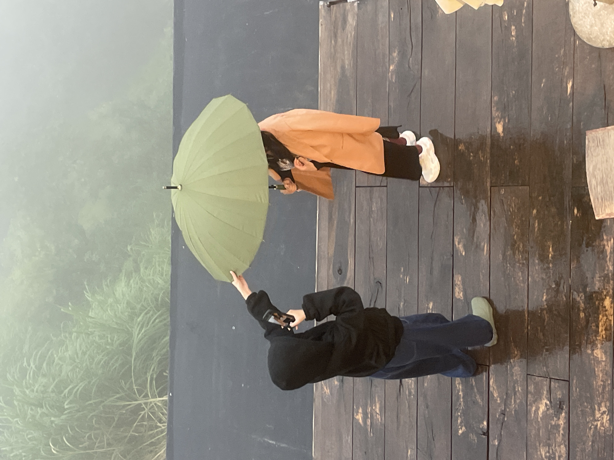 Two individuals stand on a wet wooden deck with a mountainous, misty landscape in the background. The person on the left, wearing a black hooded jacket and blue pants, holds a smartphone to take a photo. The person on the right, wearing an orange jacket over dark clothing and white sneakers, holds a large green umbrella over both of them. The scene appears to be at a cafe or viewpoint in Da Lat, Vietnam, possibly at a location like the Datanla Waterfall area which features similar wooden decking and mist-covered vegetation.