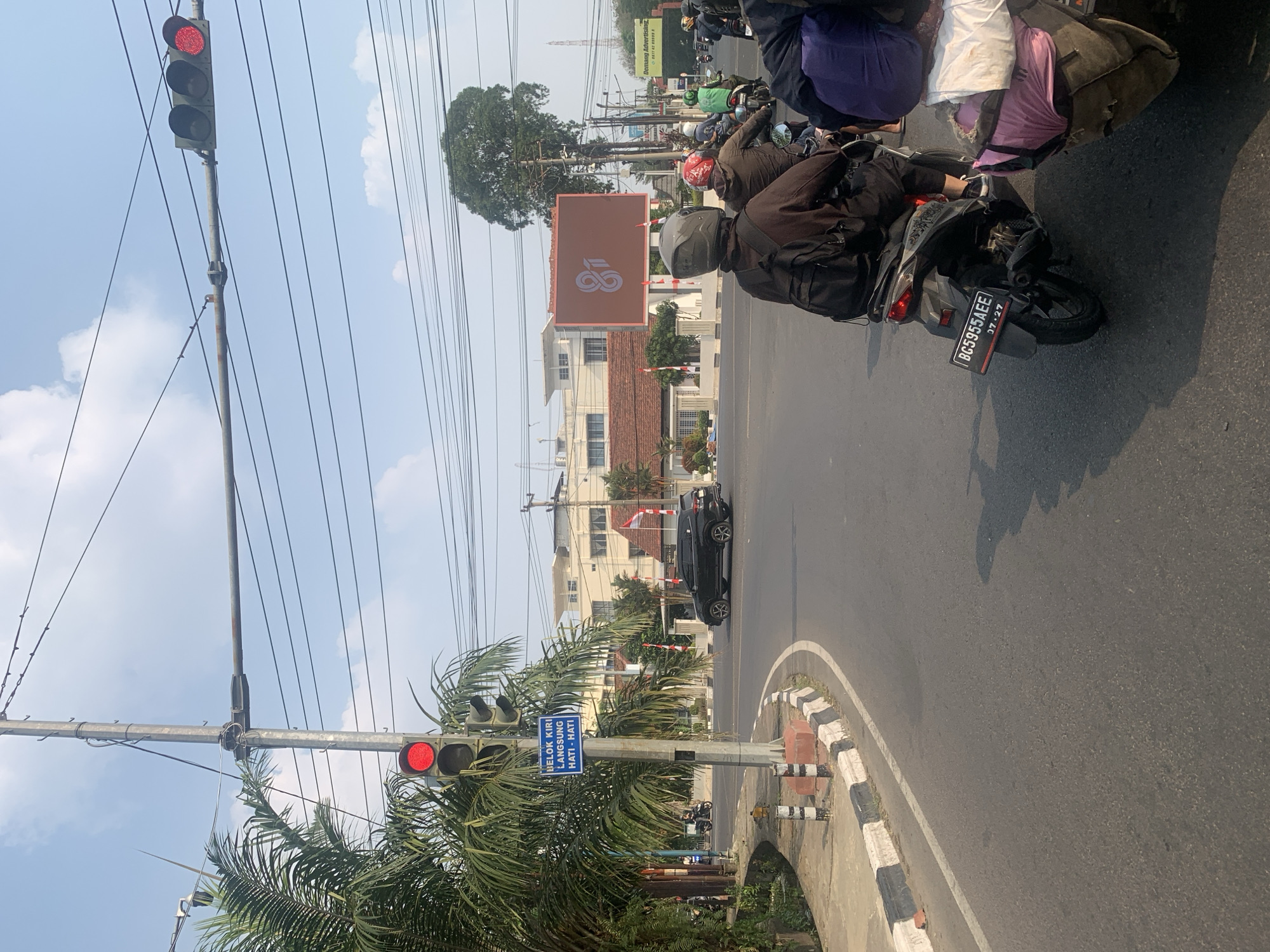 The image shows a street scene in Palembang, Indonesia, likely an intersection with a traffic light. A traffic light is visible, displaying a red signal. On the side of the road, there is a blue sign with white text that appears to be a traffic instruction. Palm trees and other vegetation are present along the sidewalk. Several motorcycles are on the road, with riders wearing helmets. One motorcycle has a license plate that reads "BG5955AEE". In the background, there are buildings, including one with a large digital billboard displaying a logo. The sky is clear with some clouds. The road is paved and appears to be in good condition. The overall scene depicts a typical urban environment with traffic and daily activity.