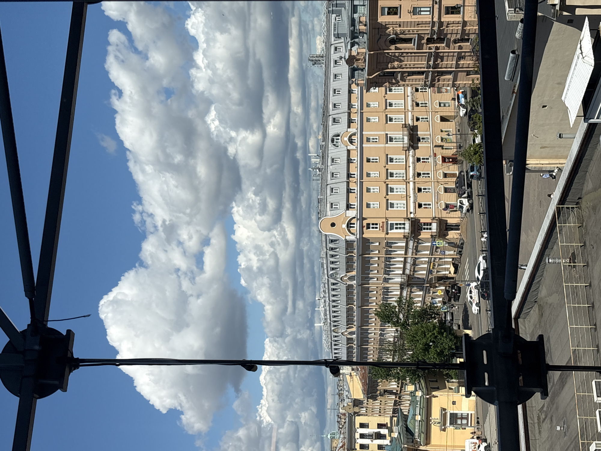 The image shows a view of Saint Petersburg, Russia, on a partly cloudy day. In the foreground, a black metal structure obstructs the view, with a black cable hanging vertically. Beyond this, a street scene unfolds with several buildings lining the road. The central building is a light yellow, multi-story structure with numerous windows and a mansard roof. To the left and right of this building are other architectural structures, some with ornate details. Cars are visible on the street, including a white police car with markings. Pedestrians can be seen walking on the sidewalk and crossing the street. The sky is a vibrant blue with large, fluffy white clouds. The overall impression is of a sunny day in a European city.