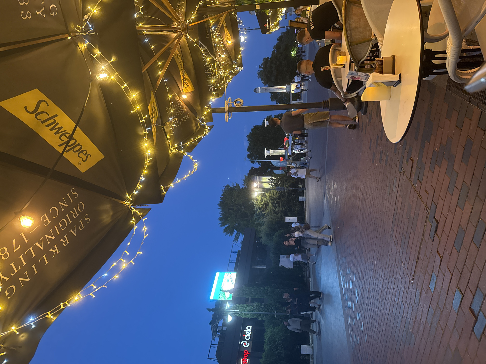 The image depicts an outdoor seating area of a cafe or restaurant in Varna, Bulgaria, at dusk. Large black umbrellas, adorned with string lights, cover the tables. One umbrella prominently displays the Schweppes logo. Several people are visible in the scene: a group walking on the paved walkway in the background, a man in a grey t-shirt and shorts walking away from the camera, and two men seated at a table in the foreground, facing away from the viewer. One of the seated men is wearing a black t-shirt with "BALENCIAGA" visible on the back. Another person, a woman, is walking in the background with a companion, wearing ripped jeans and a patterned top. The walkway is paved with brick-like material, with some darker rectangular inlays. In the distance, a large digital billboard is visible, and architectural structures with columns can be seen. The overall atmosphere suggests a lively evening setting.
