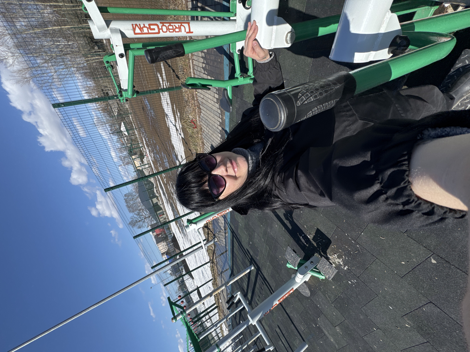 A woman wearing sunglasses and a black jacket is posing for a photo on a piece of outdoor exercise equipment labeled "TURBO GYM". She is holding a dark-colored bottle. The equipment is white and green, and it's situated on a black, textured surface. In the background, there is a chain-link fence, patches of snow, bare trees, and buildings. The sky is blue with some white clouds. The scene appears to be an outdoor exercise area in Zav'yalovo, Russia.