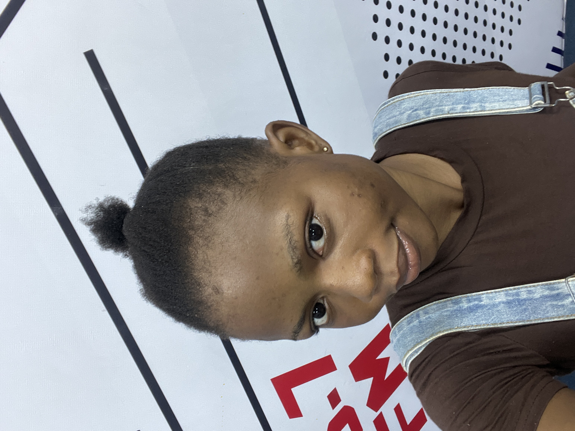 A close-up shot focuses on a person with dark hair pulled into a small bun, and dark eyes looking directly at the camera. The person wears a brown shirt with light denim straps. The background is a white wall with black stripes and a dotted pattern. Red letter outlines are partially visible in the lower part of the frame. There is no definitive information about the location being Lagos, Nigeria.