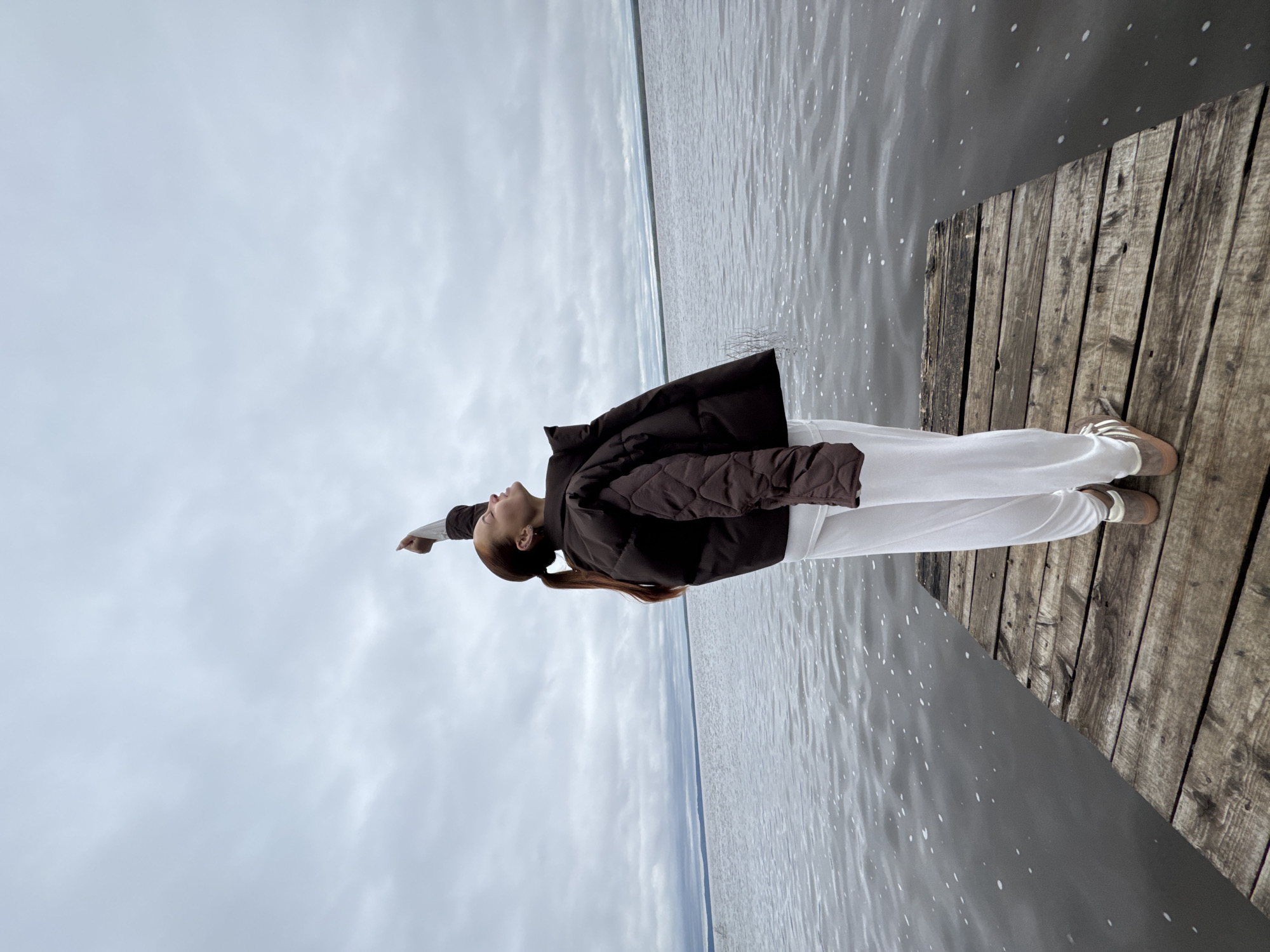 A young woman stands on a wooden pier next to water, her head turned upwards and her right arm extended. She wears a brown quilted jacket, white pants, and light-colored shoes. The sky is overcast. The location is possibly in Chastyye, Russia, based on the prompt.