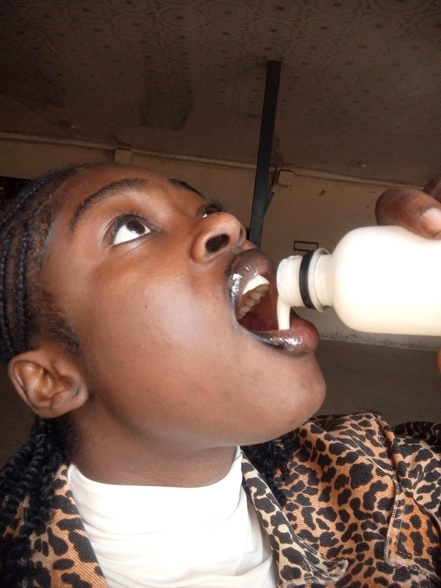 A close-up, low-angle shot shows a person drinking from a white bottle. The person is looking up and to the left, with their mouth open to receive the liquid. Their lips are glossy and their teeth are visible. The bottle, held by a hand, is pouring a white liquid into their mouth. The person is wearing a white shirt and a jacket with a leopard print. The background is out of focus, showing a brown, textured ceiling and a tan wall with a dark vertical beam. The scene appears to be indoors.
