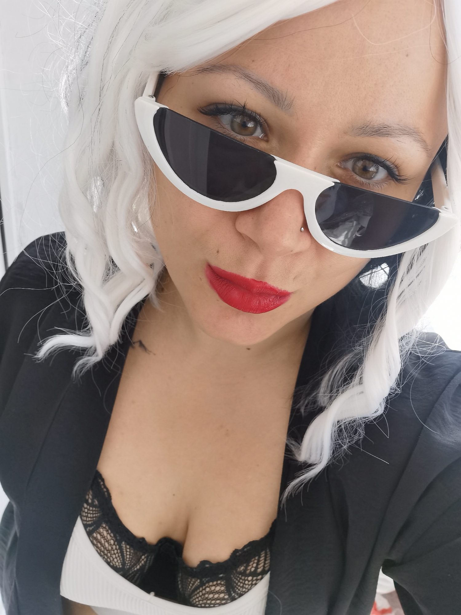 A close-up portrait features a person, likely a woman, with fair skin and dark brown eyes. She wears distinctive white-framed sunglasses with dark lenses, which sit high on her face. Her hair is bright white and styled in soft waves, possibly a wig. A small silver philtrum piercing is visible above her upper lip, and she has bright red lipstick.
She is dressed in a black blazer or jacket, under which a black lace bra is visible over a white ribbed top. The person's gaze is directed confidently towards the camera. Her eyelashes appear long and dark. On her right shoulder/neck area, a small dark mark or tattoo can be seen. The background is out of focus and light-colored, suggesting an indoor setting with bright, even lighting. There is no visible text in the image. No specific elements within the image directly indicate the stated location of Chaikovsky, Russia.