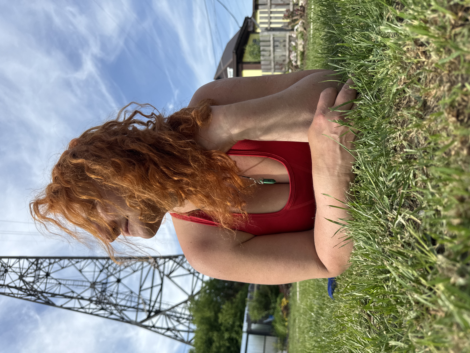 A fair-skinned woman with long, curly auburn hair is lying on a lawn of green grass. She is wearing a red top. Her arms are crossed and folded in front of her, and she is looking to the left. In the background, there is a partially visible utility pole, a building, and a blue sky with clouds. The photograph was taken in the city of Chaikovsky, Russia.
