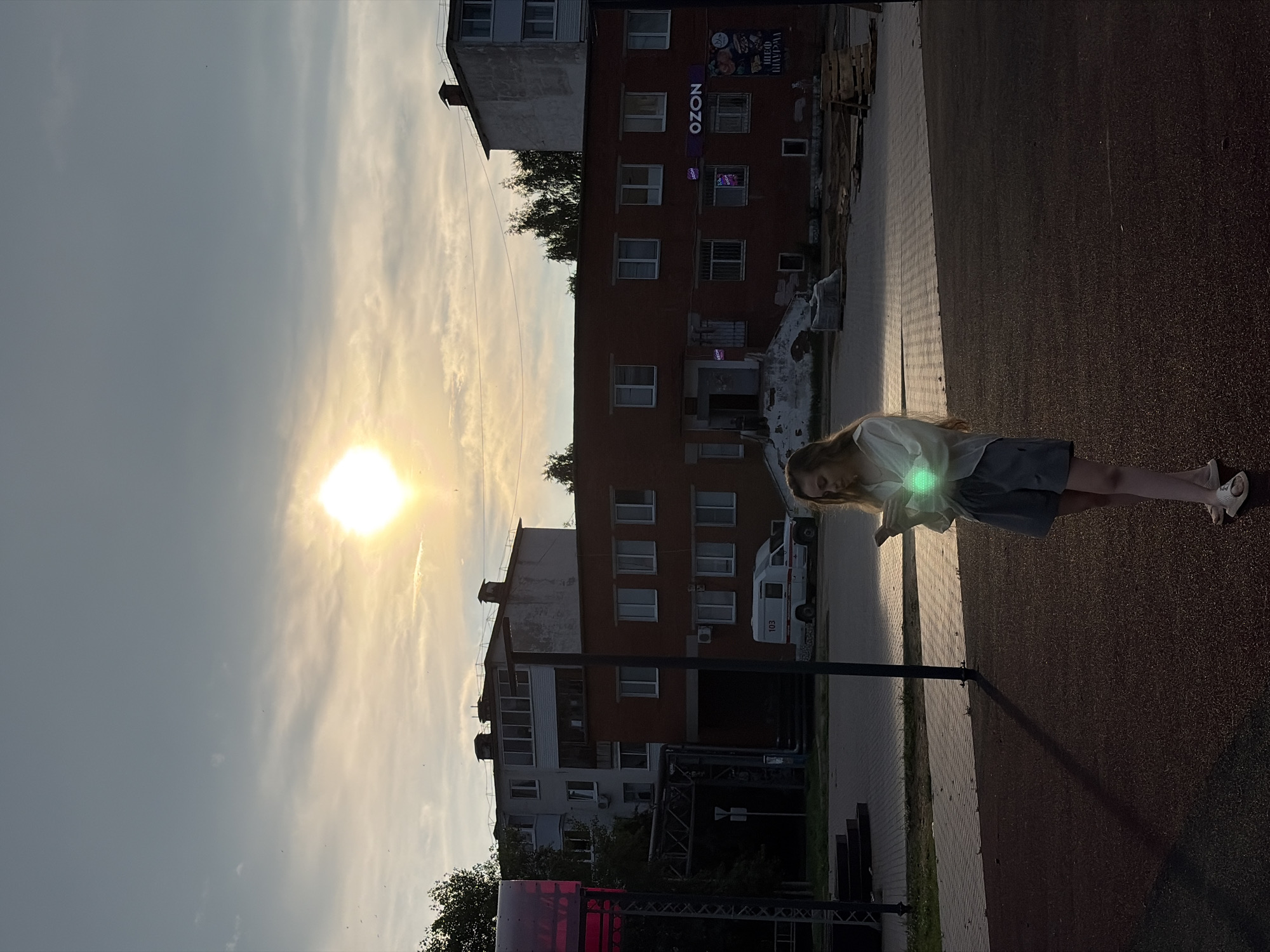 The image depicts a young woman standing in an outdoor area, possibly a park or a public square, with a bright sun low in the sky, casting long shadows. The woman is wearing a light-colored shirt and shorts and appears to be holding a smartphone, which is emitting a green glow. In the background, there are several brick buildings, one of which has "OZON" signage. A white vehicle with "103" markings is parked in front of one of the buildings. The overall environment suggests an urban setting, possibly in Noviy, Russia, given the context provided. The lighting indicates it is late afternoon or early evening.