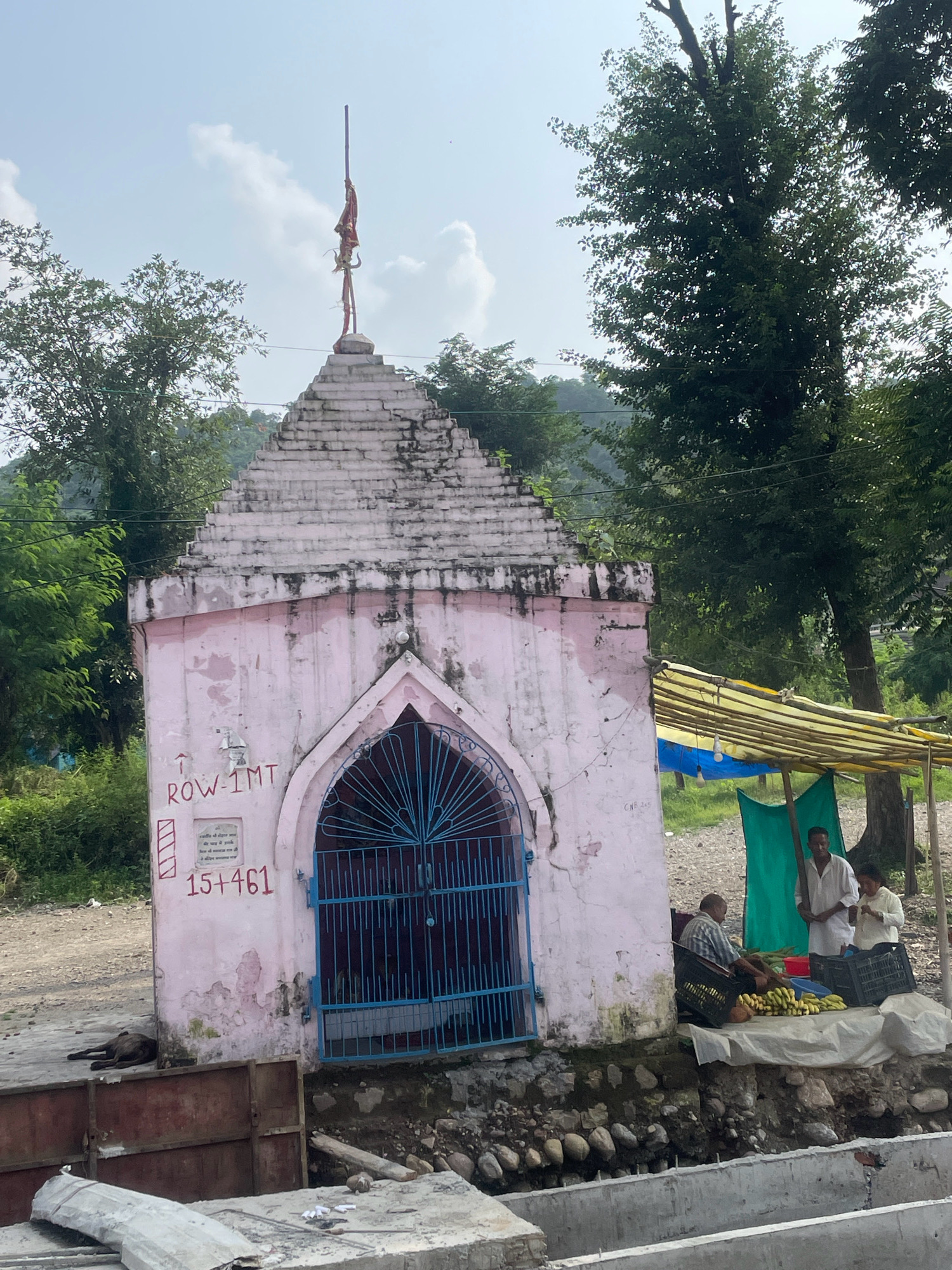 This image captures a small, light pink, weathered temple in Raja ka Bagh, India. The temple has a tiered, pyramid-shaped roof topped with a flagpole bearing a flag. In front of the temple is a blue metal gate. To the right of the temple, a makeshift stall is set up with a yellow and blue tarp overhead. Two men and a woman are present at the stall. One man is sitting and appears to be selling bananas and coconuts, which are displayed in crates and on a white cloth. Another man and the woman are standing, interacting with the produce. The ground around the stall is covered with pebbles and some vegetation. A dog lies on the ground to the left of the temple. The background consists of lush green trees and a hilly landscape under a partly cloudy sky. Construction materials, including concrete slabs and metal sheets, are visible in the foreground.