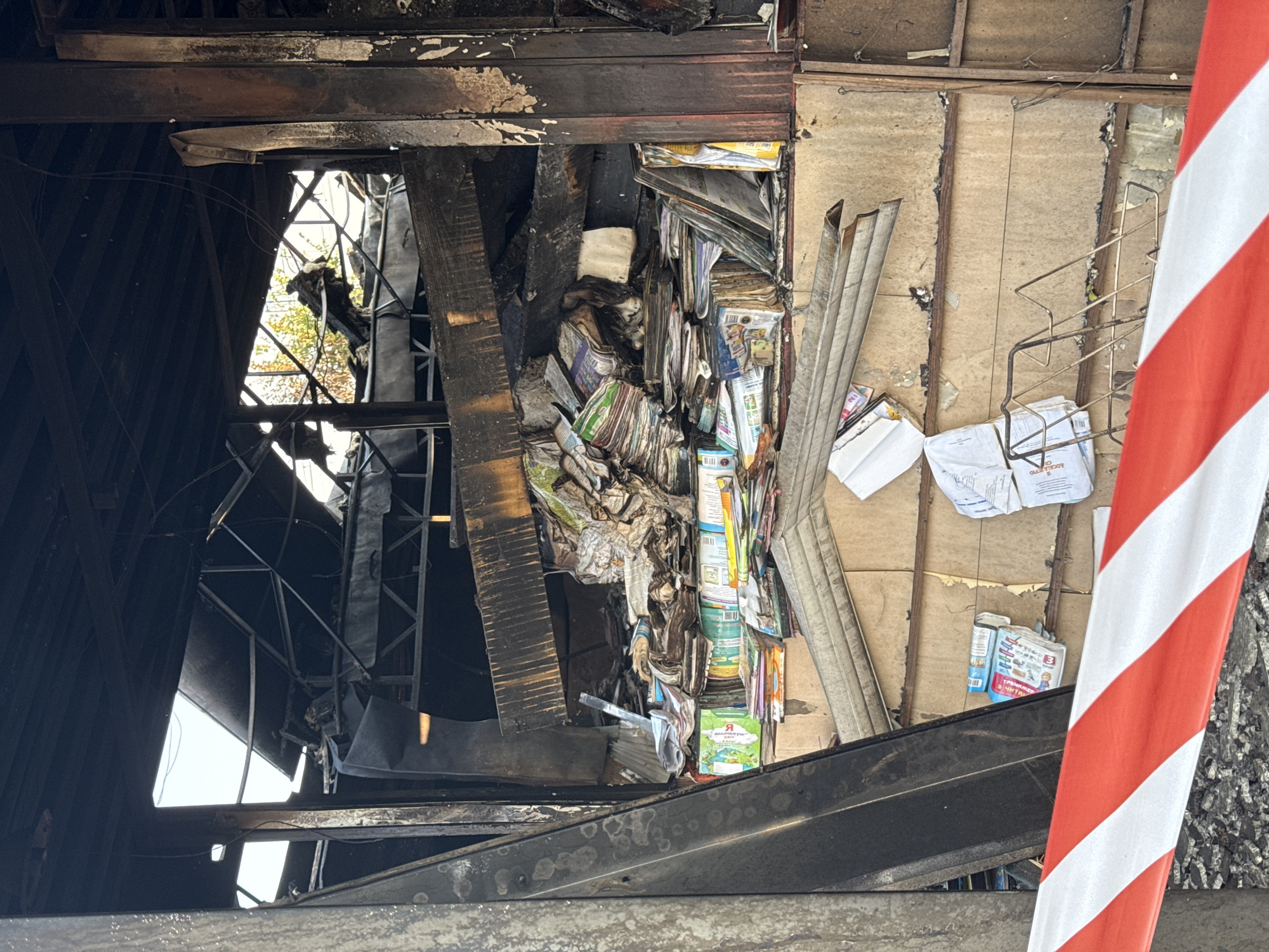 This is a medium shot, eye-level, vertical photograph showing the interior of a building that appears to have been damaged by fire. The structure is partially collapsed, with exposed beams and charred debris. In the foreground, a red and white striped caution tape stretches across the bottom of the frame. The midground is dominated by stacks of books and papers, some of which are scattered and appear water damaged. The books are of various sizes and colors, with some exhibiting faded covers and warped pages. Behind the books, the wall of the building is visible, made of textured panels and showing signs of soot and discoloration. In the background, the open ceiling reveals more structural damage and a glimpse of the sky. The overall impression is one of destruction and disarray. The provided context indicates this scene is located in Kyiv, Ukraine.