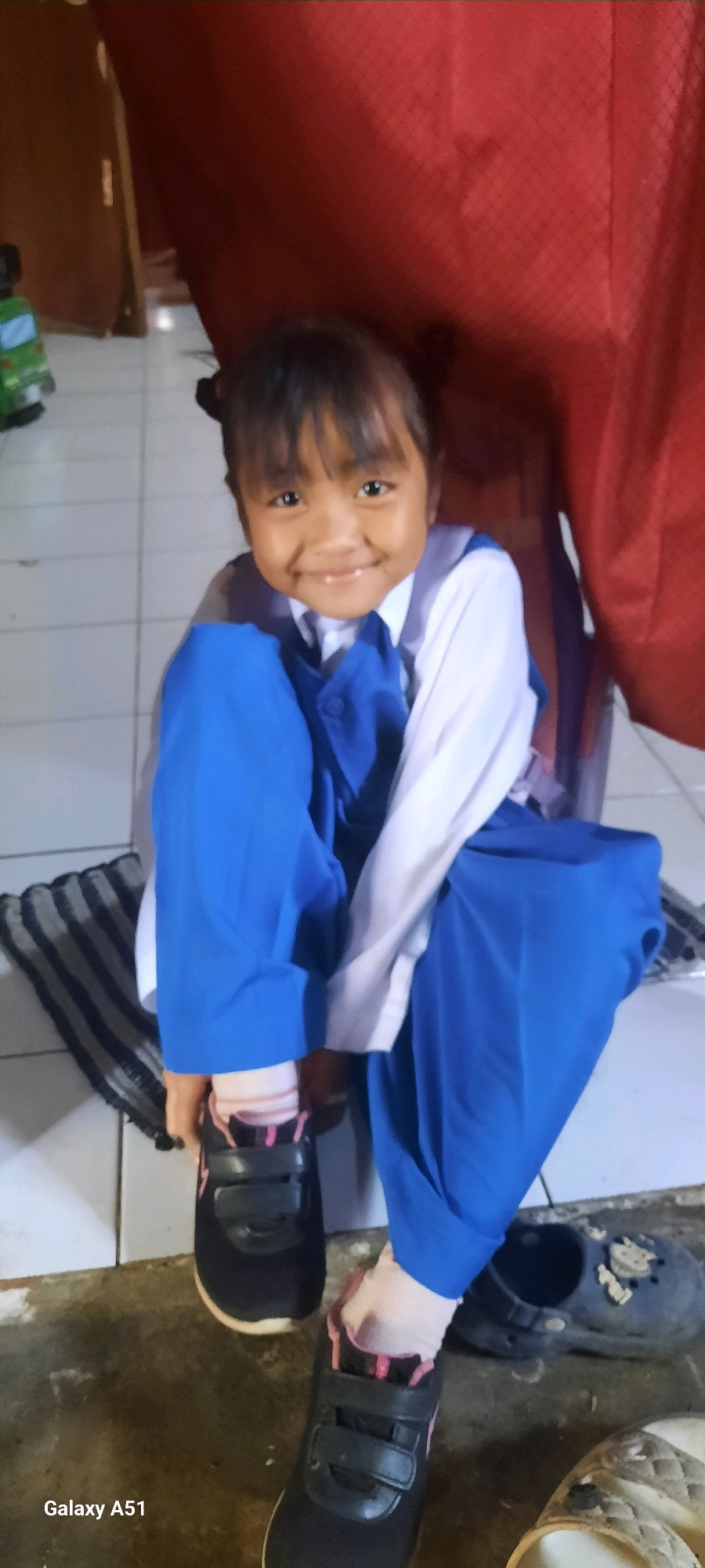 In Ploso, Indonesia, a young girl wearing a white shirt, blue vest, and blue pants is seated on a tiled floor. She is in the process of putting on a black shoe with pink accents and velcro straps, while wearing striped socks with pink trim. Beside her, another pair of black clogs and a light-colored sandal are visible. The background consists of a red curtain and a brown partition.