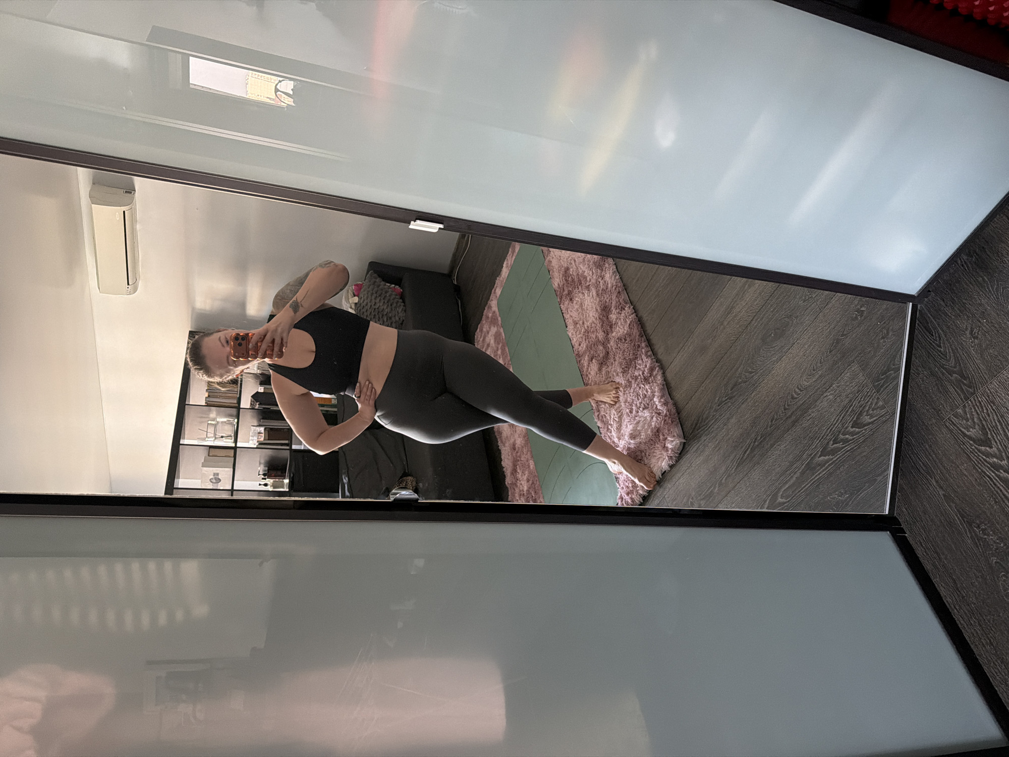 An adult female is depicted in a full-length mirror selfie taken indoors within an apartment room in Moscow, Russia. She stands barefoot on a pink shag rug, positioned with her left hand on her hip and her body slightly angled, while holding an orange-cased smartphone with both hands to capture the image. She wears a black sports bra and dark grey leggings, and visible tattoos are present on her left arm. The mirror is framed in black and appears to be part of a sliding door system.
The reflection in the mirror reveals the interior environment: dark wood-grain flooring, a folded green yoga mat placed on the pink rug, a dark grey couch or armchair, and a black open-shelf unit displaying various objects including diffusers, bottles, and books. A white wall-mounted Carrier air conditioning unit is installed high on the wall. A window reflection in the upper right portion of the mirror shows an external view of a multi-story building with a light-colored facade, indicative of an urban setting under an overcast sky.