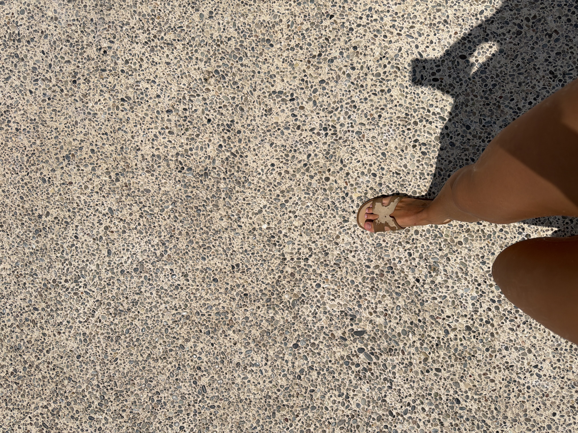 The image shows a person's legs and feet walking on a gravelly path. The person is wearing a sandal on their right foot. The shadow of the person is visible on the path, indicating a sunny day. The environment is an outdoor setting, possibly a park or a beach promenade in Oroklini, Cyprus. The path is made of small, light-colored pebbles embedded in concrete. The person appears to be walking towards the bottom of the frame.