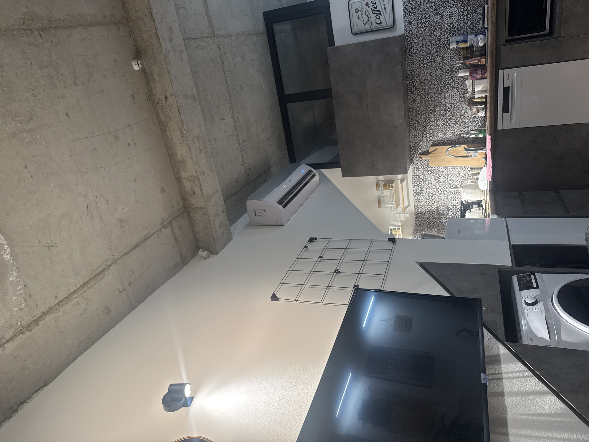 The image depicts an interior scene, likely a modern apartment kitchen and living area. In the foreground on the left, a large black television is mounted on a wall. Below the television, a white washing machine is visible. The walls are white, and the ceiling is constructed of exposed concrete. A black wall-mounted light fixture is on the left wall, and a white air conditioning unit is mounted on the wall towards the upper right.
The background reveals a kitchen area with dark cabinets, a patterned backsplash, and a wooden countertop. Various kitchen appliances and items, including a refrigerator, kettles, containers, and a microwave, are present on the countertop and shelves. A wooden cutting board with a faucet is positioned over the sink area. A framed "Coffee" sign hangs on the wall above the kitchen counter. The overall aesthetic appears to be minimalist and industrial, with a mix of textures like concrete, tile, and wood. There are no people visible in the image.