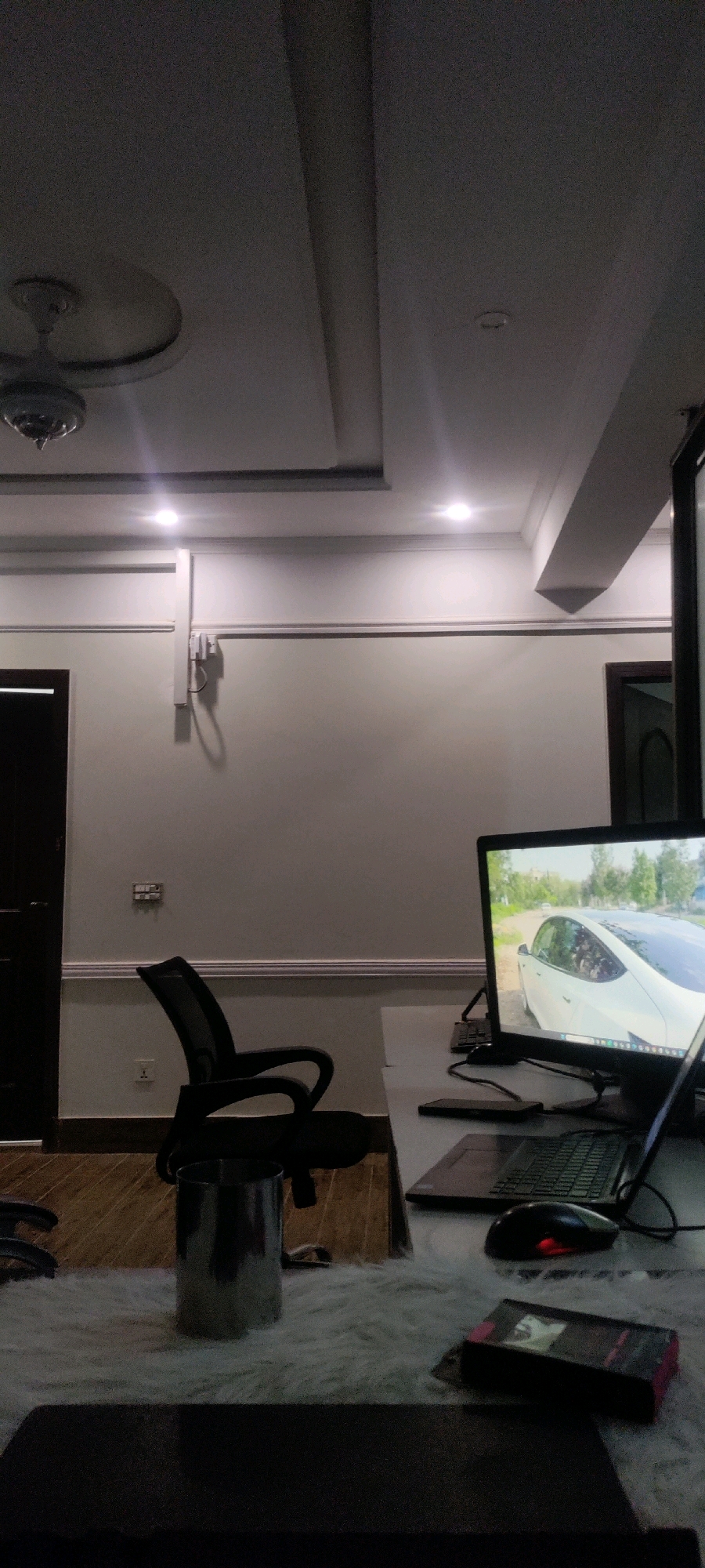 This is an indoor scene, likely an office or home office, located in Khana Dak, Pakistan. The room is dimly lit, with recessed ceiling lights casting a soft glow. A ceiling fan is visible in the upper left portion of the image. The walls are painted a light neutral color and feature white molding near the ceiling and floor. A dark wooden door is present on the left side of the frame, and another partially visible dark wooden door is on the right.
In the center of the image, a black office chair with a mesh back is positioned. In front of the chair is a desk with a white surface. On the desk, a computer monitor displays an image of a white car. A laptop is open next to the monitor, with a black computer mouse with a red illuminated scroll wheel positioned in front of it. To the left of the laptop, a dark smartphone or similar rectangular electronic device is visible. Further to the left, a silver cylindrical container sits on a fluffy white rug.
In the foreground, a dark rectangular object, possibly a laptop or a large notebook, rests on a dark surface. The overall impression is of a quiet workspace, possibly at dusk or during nighttime hours.