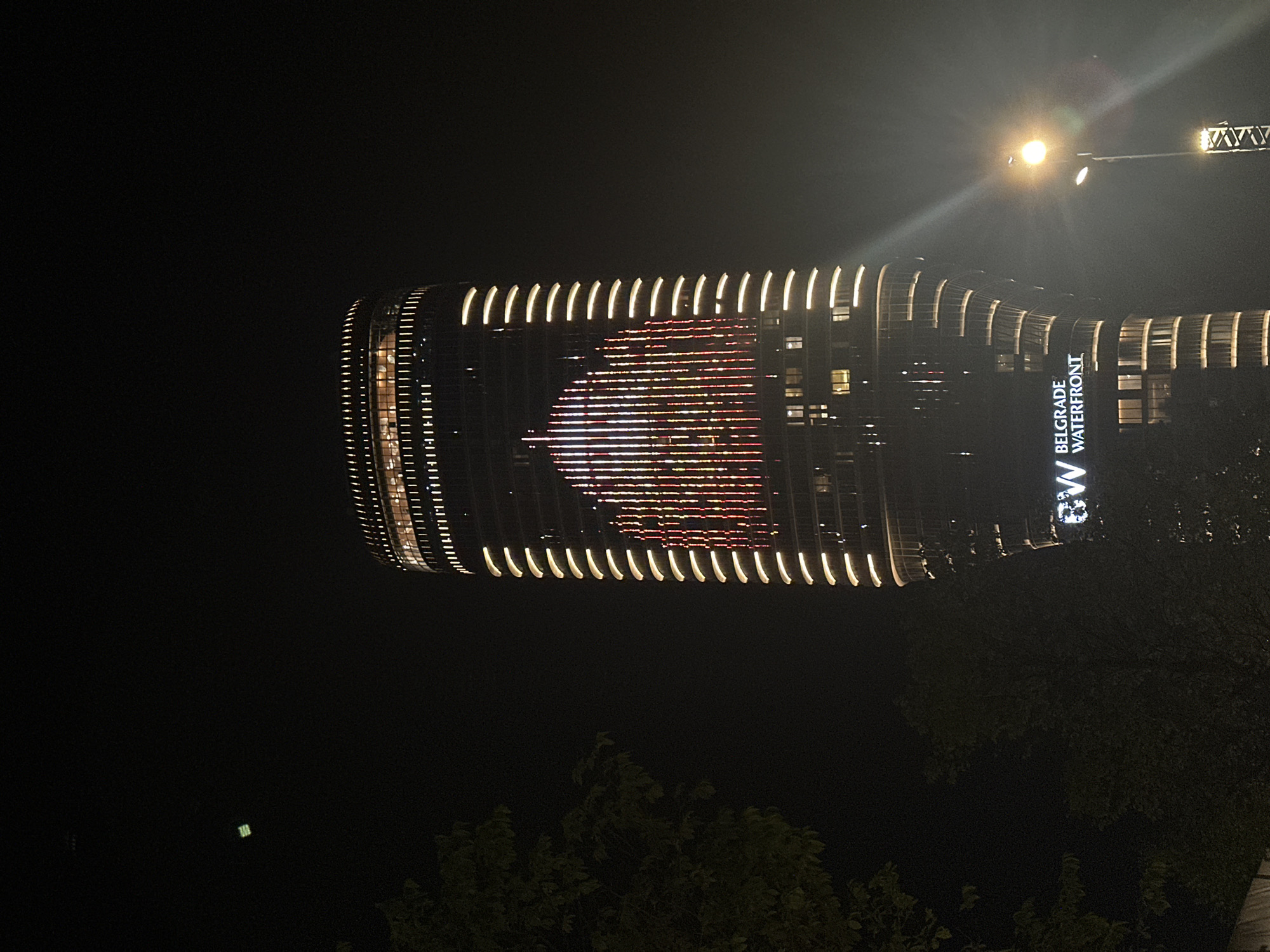 At night, a tall cylindrical building illuminated with horizontal lines of light, and displaying a pattern resembling a stylized heart in the center, stands prominently. The building is part of the Belgrade Waterfront development, as indicated by the prominent "BELGRADE WATERFRONT" signage. A streetlamp is visible on the upper right, casting a bright light. A few trees are visible in the lower portion of the image.