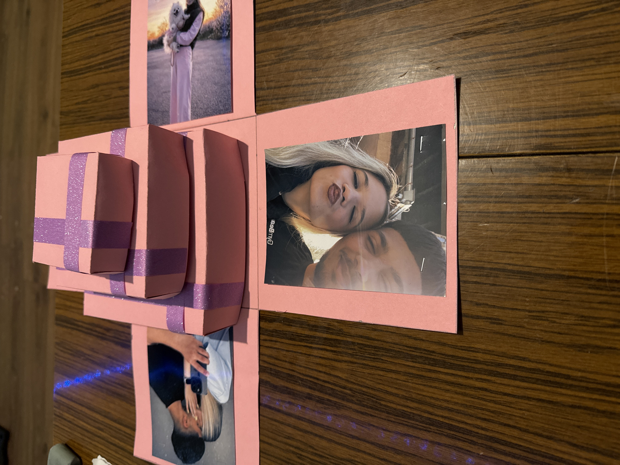An open, pink explosion box, a creative photo album and gift, is displayed flat on a dark brown wooden surface. The box is made of pink cardstock and features multiple flaps that unfold, each adorned with a photograph.
In the top section, one photo shows a person in light-colored clothing holding a small white dog outdoors, illuminated by the warm glow of what appears to be sunrise or sunset. To the right, a vertical photo (rotated 90 degrees clockwise) captures a woman with blonde hair playfully pursing her lips, while a man with short dark hair rests his head near her, both looking towards the camera. Partial text "®wgæ" is visible on the woman's black garment. The bottom section presents another vertical photo (rotated 90 degrees clockwise) of a man and a woman in a tender embrace.
At the center of the explosion box, three smaller, pink gift boxes are stacked, each tied with a shimmering purple ribbon. The overall arrangement suggests a thoughtful and personalized gift. The wooden surface beneath shows distinct grain patterns.
The items are presented in an indoor setting, and the emotional tone conveyed by the photographs is one of affection and happiness. The location context provided is Yana, Bulgaria, though no visual cues in the image directly confirm this.