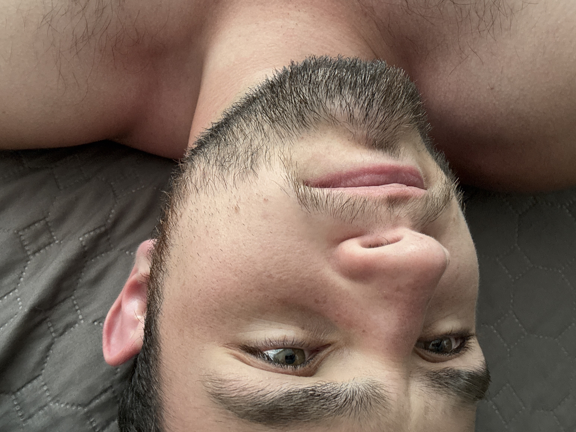 A close-up, upside-down view of a man's face is presented. He has short dark hair, a full beard, and fair skin. His eyes are open, and he appears to be lying on a gray textured surface, possibly a bed. His shoulders are visible in the background. There is no information in the image to suggest a location in the city of San Bernardino Tlaxcalancingo, Mexico.