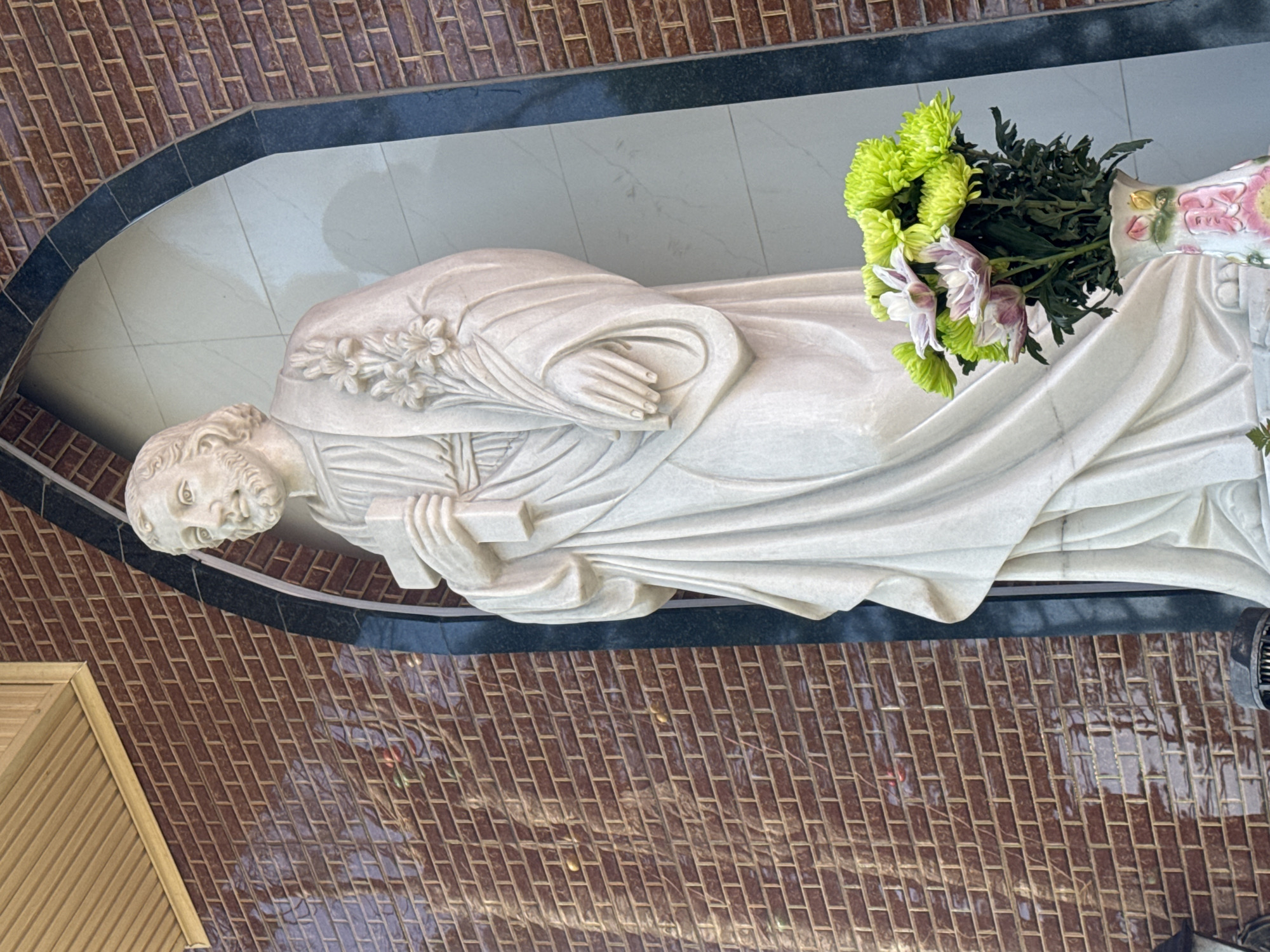 A white marble statue of a male figure, likely religious, stands within a dark arched niche against a brick wall. The figure is depicted from the waist up, holding a cross in its right hand and a bouquet of flowers in its left. The statue has a beard, flowing hair, and wears a draped robe adorned with carved flowers on its shoulder. In the foreground, a vase containing a bouquet of green and pink chrysanthemums is placed at the base of the statue. The brick wall behind the statue is red and appears to be glazed, reflecting light. The niche itself is lined with lighter colored tiles. Above the niche, a wooden soffit with horizontal planks is visible. The overall setting suggests an outdoor or semi-outdoor religious or memorial location, possibly within a church or cemetery. The image was taken in Tuý Sơn, Vietnam.