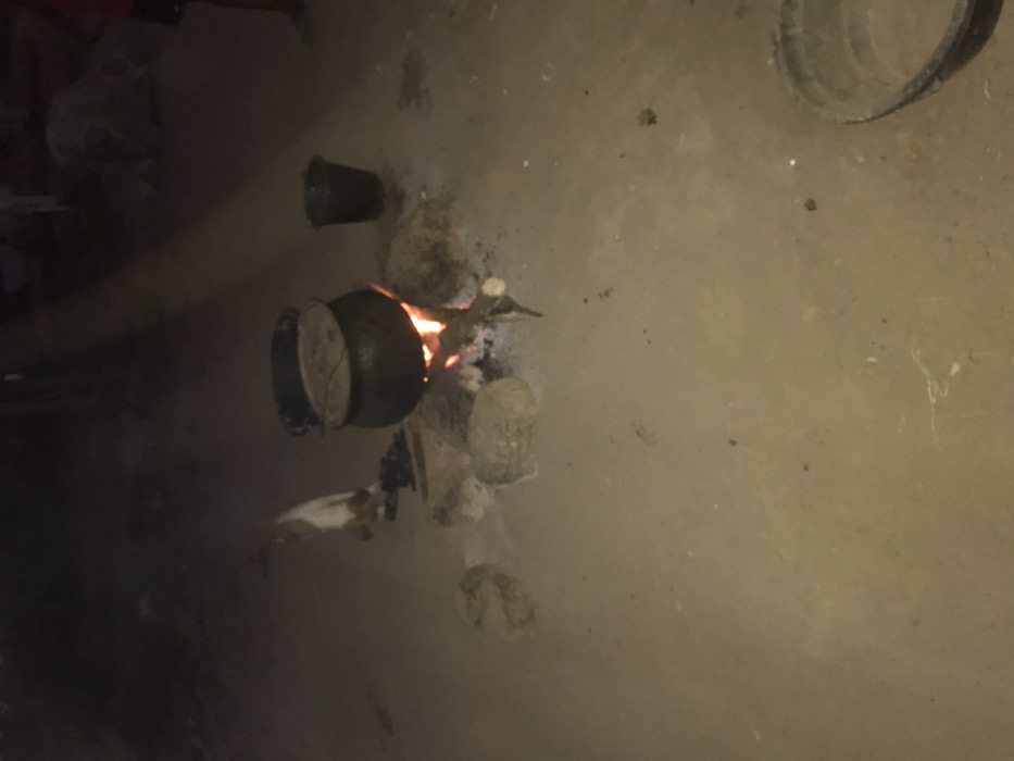The image depicts a dark, likely nighttime, scene showcasing an outdoor cooking arrangement. A large, dark pot with a lid is centrally placed over a bright, open fire, fueled by burning wood. Several rocks are strategically positioned around the fire, serving as a makeshift stove to support the pot. The fire casts an orange glow, illuminating the immediate area. A small animal, resembling a cat or a small dog, is visible on the ground near the fire. To the right of the main cooking area, a smaller, dark bucket or pot is present. The ground beneath is light brown, appearing to be dirt or sand. In the upper right corner, a large, dark cylindrical object, possibly another bucket or a tire, is partially visible. This traditional cooking method suggests a rural or undeveloped setting, aligning with the provided location context of Ago-Abo, Nigeria. No people are visible, and there is no discernible text.