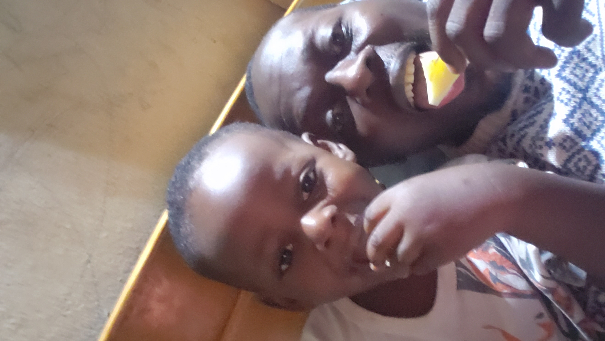 In Sabon Tasha, Nigeria, two individuals, an adult male and a child, are captured in a close-up, selfie-style photograph. The adult male, positioned on the right, has his mouth open and is holding a slice of yellow fruit, possibly lemon, to his lips. He is wearing a patterned sweater. To his left, a child is also visible, facing the camera with a piece of food near their mouth. The background appears to be an indoor setting with a textured wall and a yellow-colored surface that could be furniture. The lighting suggests an indoor environment, possibly at home.