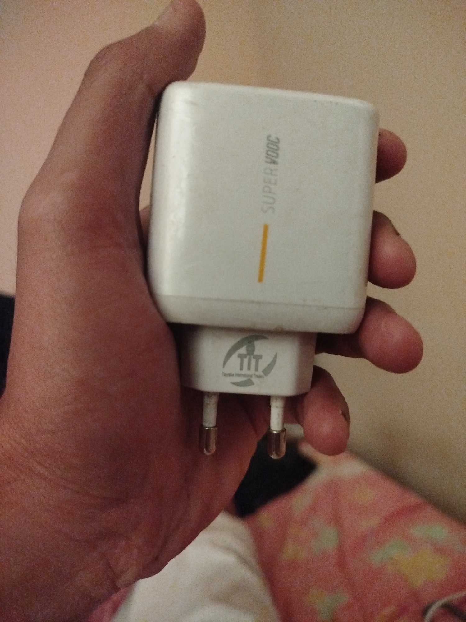 A person's hand is holding a white fast charger. The charger has "SUPER VOOC" printed vertically on its side in white text. Below the main charger unit is a smaller adapter with a logo that includes the letters "TIT" in a stylized font. The charger has two metal prongs extended downwards. The background is out of focus, showing a light-colored wall and a pink patterned fabric, possibly a blanket or bedsheet. The lighting suggests an indoor setting, possibly during the day or with artificial light. The image is a close-up shot focusing on the charger and the hand holding it.