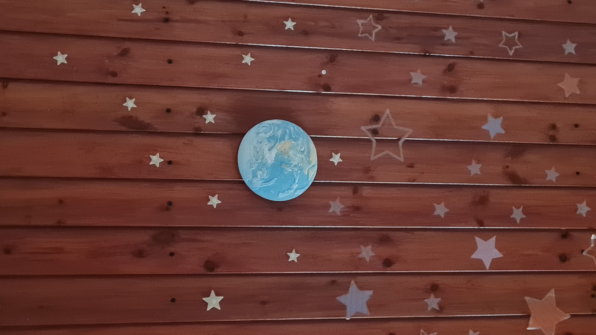 A close-up shot reveals a wooden wall, comprised of horizontal planks with a reddish-brown hue. Adorning the wall are various star-shaped decorations, some solid and some just outlines. In the center, there's a circular object with a blue and white marbled pattern, resembling a depiction of the Earth. The scene appears to be indoors, with the decorations possibly creating a space-themed aesthetic. The location is mentioned as Lachish Regional Council, Occupied Palestine.