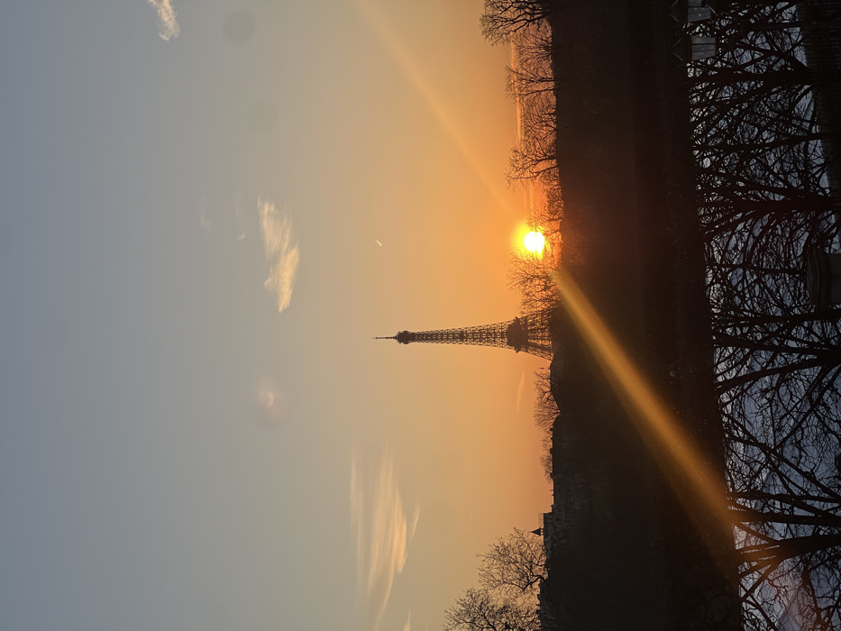 The image displays the Eiffel Tower in Paris, France, at sunset. The sky exhibits a gradient, transitioning from a light blue at the top to an orange-yellow hue near the horizon. The sun, appearing as a bright orange orb, is positioned on the right side of the frame, partially obscured by a silhouette of bare deciduous trees and distant urban structures. The Eiffel Tower stands centrally, silhouetted against the bright sunset glow. A prominent orange lens flare diagonally traverses the lower-left quadrant of the image, originating from the sun. Below the horizon, the dark silhouettes of bare trees and several distinct lampposts are reflected in a body of water, identified as the Seine River. Faint, scattered cloud formations are present in the upper sky, and a slender crescent moon is visible in the upper right quadrant. No individuals or discernible actions are present within the frame.