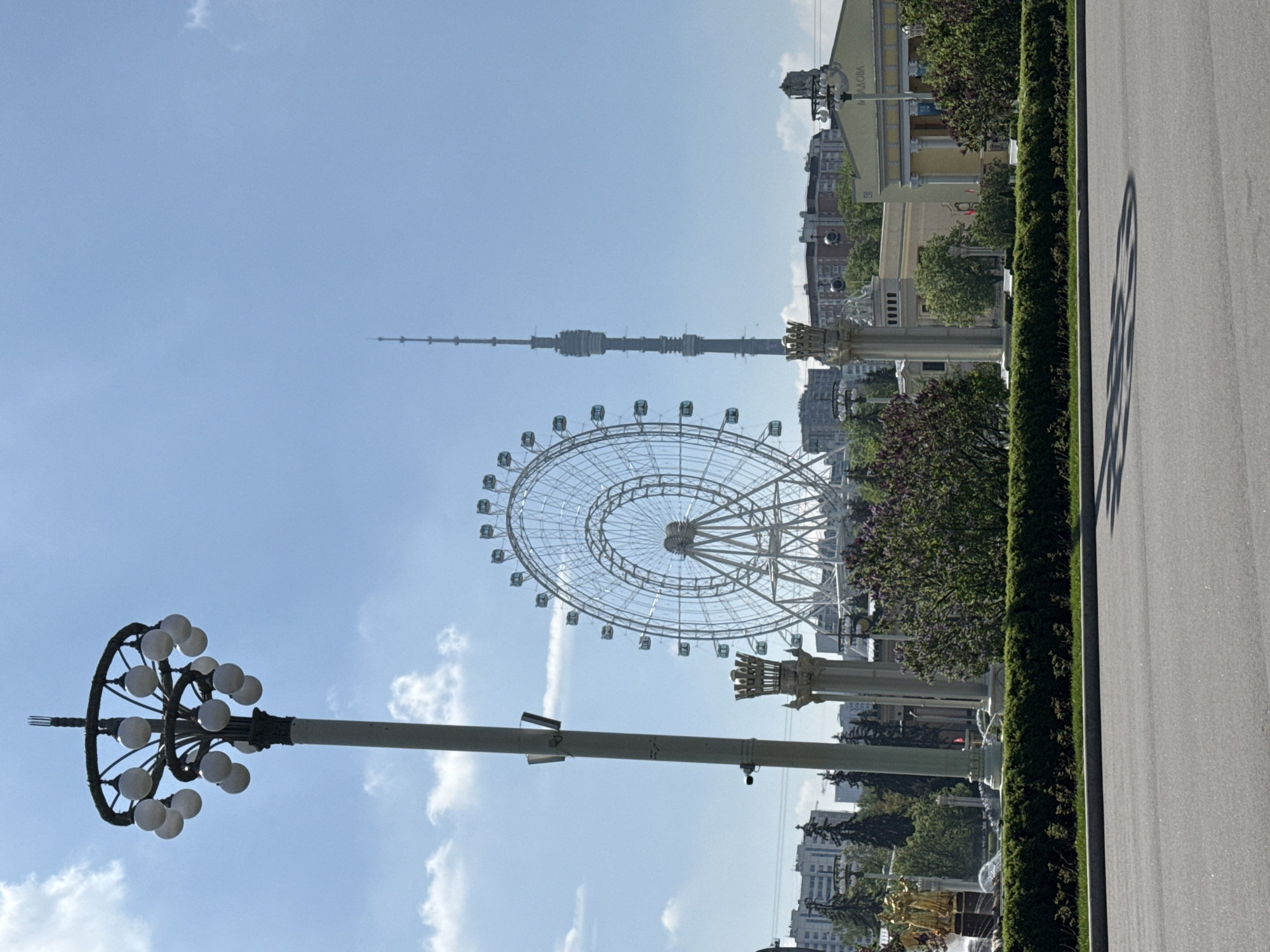 The image depicts a Ferris wheel and the Ostankino Tower in Moscow, Russia. The Ferris wheel is a large, white structure with numerous passenger cars. The Ostankino Tower is a tall, slender communications tower that is visible in the background. A lamppost with spherical lamps is in the foreground. A road and bushes are also visible, along with some buildings in the background. The sky is blue with some clouds.