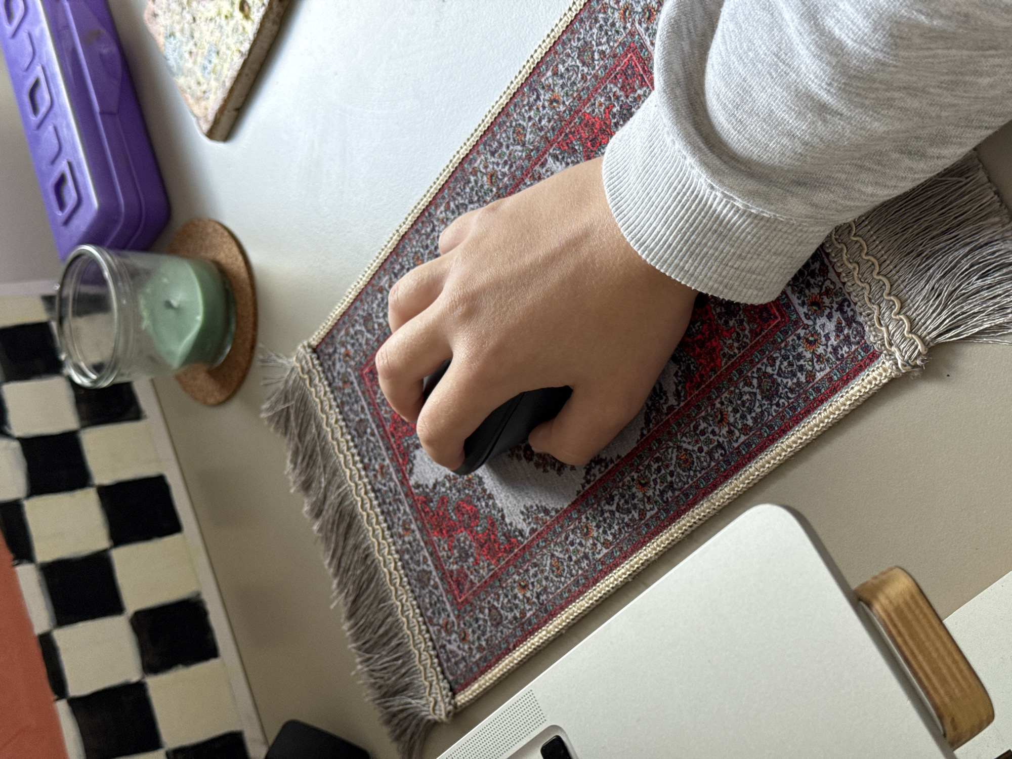 A close-up, high-angle shot captures a person's right hand operating a black computer mouse on a desk or tabletop. The hand is positioned over a decorative mousepad that resembles a miniature Persian rug, featuring intricate patterns in shades of grey, red, and subtle green, complete with fringed edges. The person wears a light grey long-sleeved top with ribbed cuffs, visible on their forearm.
Part of a silver or light-colored laptop, likely a MacBook, is visible in the bottom right corner of the frame, indicating a computer workstation. Surrounding objects on the light-colored surface include a clear glass jar with a partially melted green candle inside, resting on a round cork coaster to the left of the mousepad. A purple multi-outlet power strip or cable organizer is visible further to the left. In the upper center, a textured, possibly stone or ceramic, object with mottled patterns lies on the desk. A segment of a black and white checkered pattern, possibly a rug or mat, is also visible in the bottom left corner.
The image depicts an indoor setting, likely a home office or personal desk area, where an individual is engaged in computer-related activity. The scene conveys a sense of everyday work or leisure. The provided location context is Vozdovac Urban Municipality, Serbia. No discernible text is visible in the image.