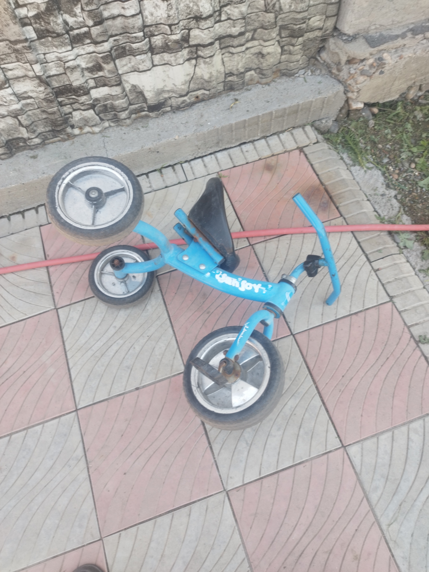 A blue tricycle is overturned on a tiled patio. The tricycle has three silver wheels and a black seat. The word "Sunjoy" is visible in white lettering on the side of the tricycle. A red hose lies on the ground next to the tricycle. The patio is made of square tiles, alternating in pink and light gray, with a curved pattern. In the background, a textured stone wall and a concrete step are visible. The scene appears to be outdoors in a residential area.