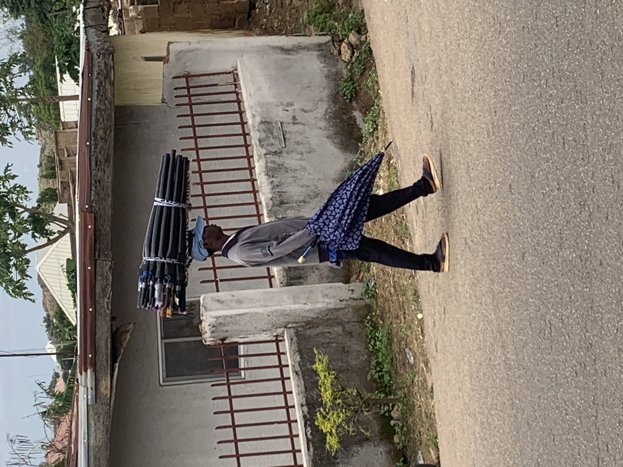 This image, captured in an urban environment within the city of Jos, Nigeria, depicts a male individual walking from left to right on an asphalt road. The man is wearing a light blue baseball cap, a grey long-sleeved collared shirt, dark trousers, and open-toed sandals. He is carrying a large, horizontally oriented bundle of black, cylindrical objects, likely folded umbrellas or thin pipes, secured with white straps, balanced on his head. In his right hand, he holds a single, folded blue umbrella with a light-colored pattern.
The immediate background on the left features a low concrete wall topped with a rusty red metal fence. Behind this fence, a light-colored building with a rectangular window is visible. A small green shrub grows at the base of the wall. To the right, another concrete wall or pillar displays discoloration and weathering, with sparse vegetation and exposed earth at its base. The distant background reveals several buildings with varied construction, including structures with rusted brown corrugated iron roofs and some with white or lighter-colored roofs. Hills or elevated terrain covered with trees and additional structures are visible further back. The sky is overcast with a uniform light grey cloud cover. The road surface appears worn asphalt.